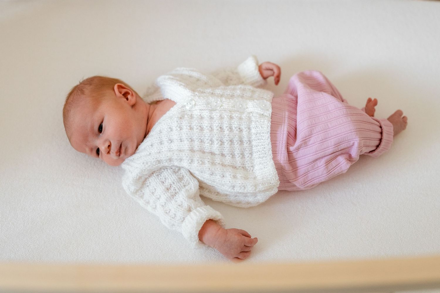 Infant in white and pink clothing resting on light colored bedding.