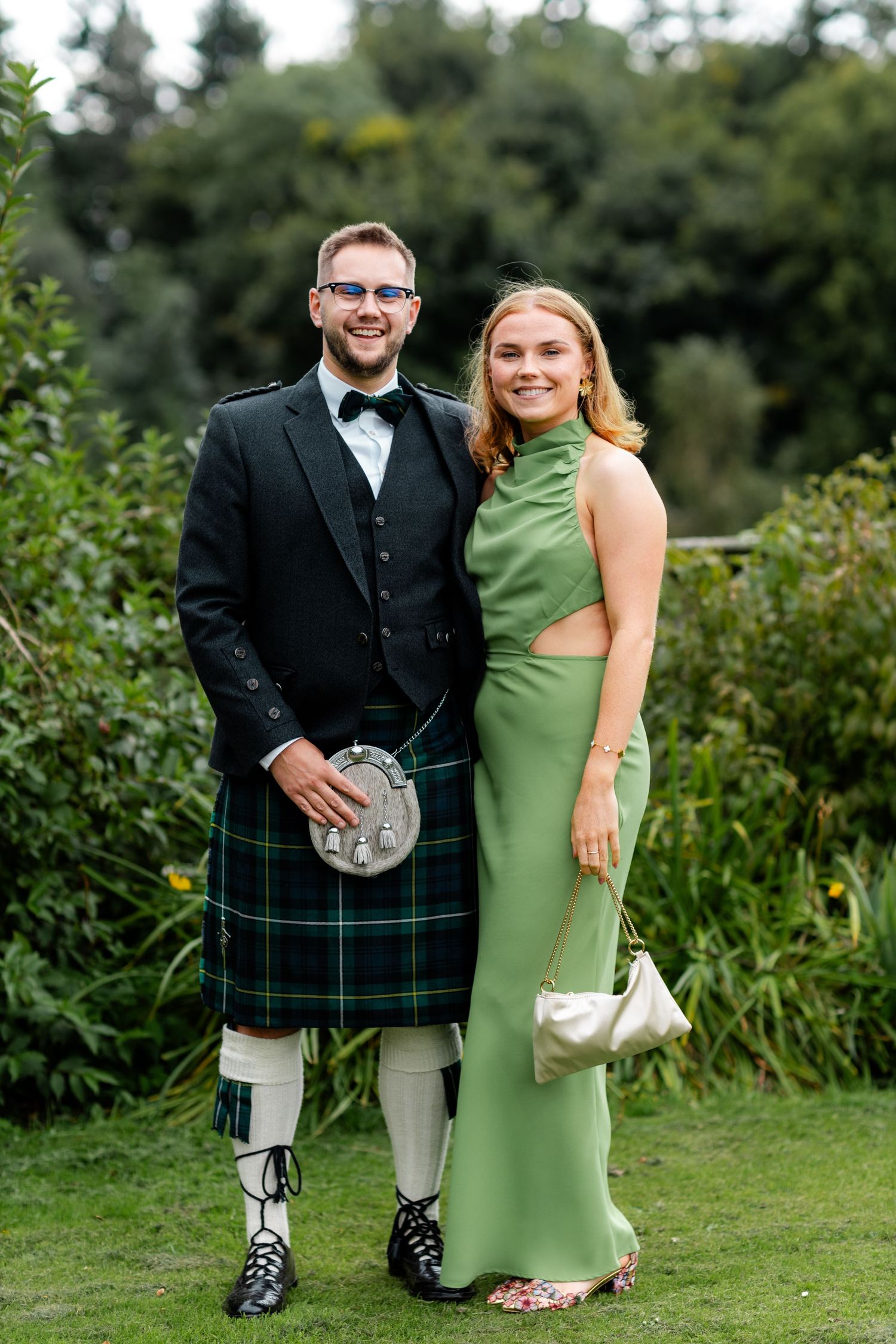 Couple posing outdoors, man in green tartan kilt and black jacket, woman in sage green gown holding white clutch.