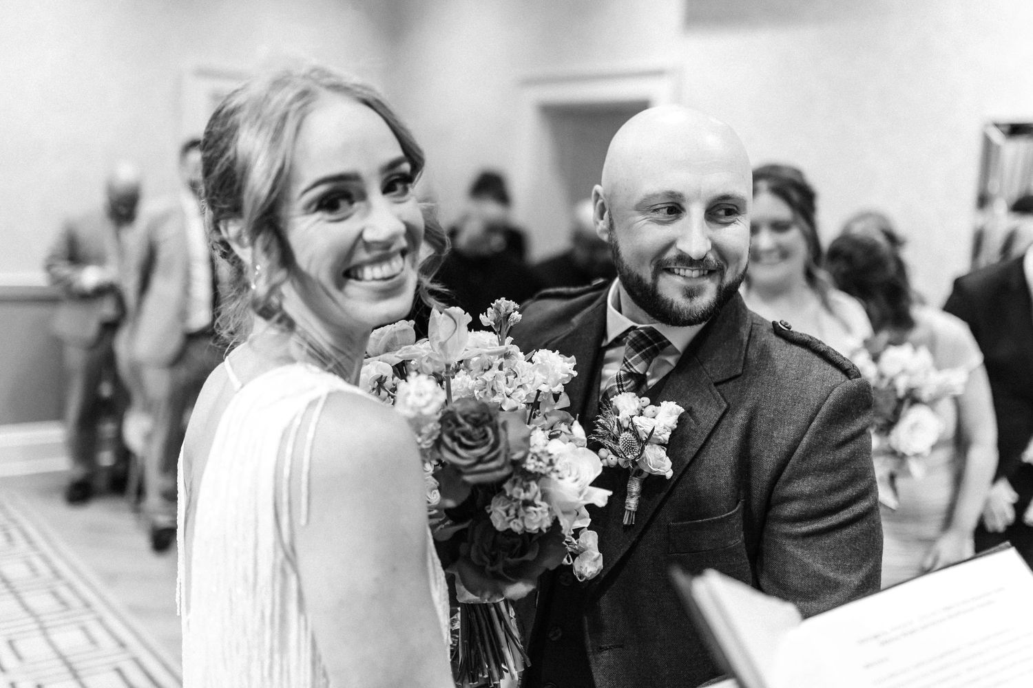 Smiling bride holding colorful bouquet stands beside bald groom in kilt at wedding ceremony in black and white.