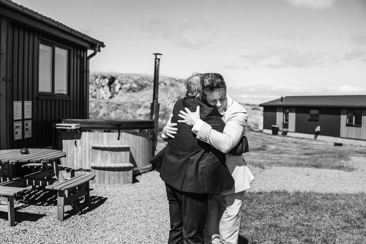 Two men share a heartfelt hug outdoors near rustic wooden cabins in a black and white wedding photo.