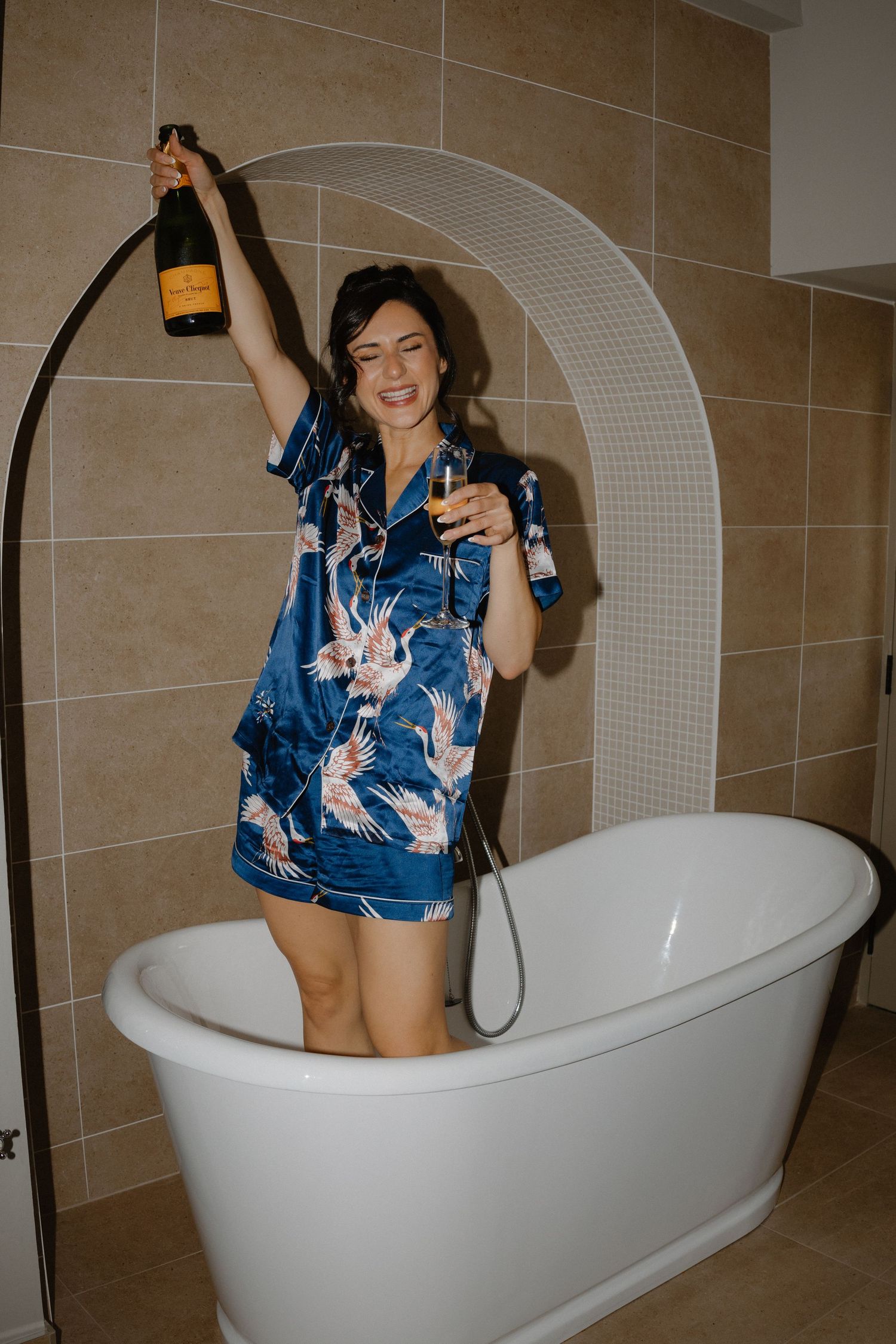 Bride in blue floral satin pajamas celebrating with Veuve Clicquot champagne while standing in a freestanding bathtub.