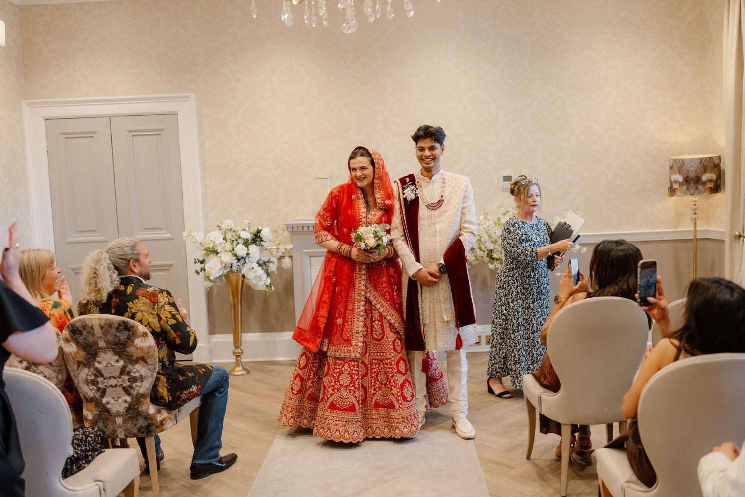 Indian bride in red lehenga and groom in white sherwani walk down the aisle at an elegant wedding ceremony.
