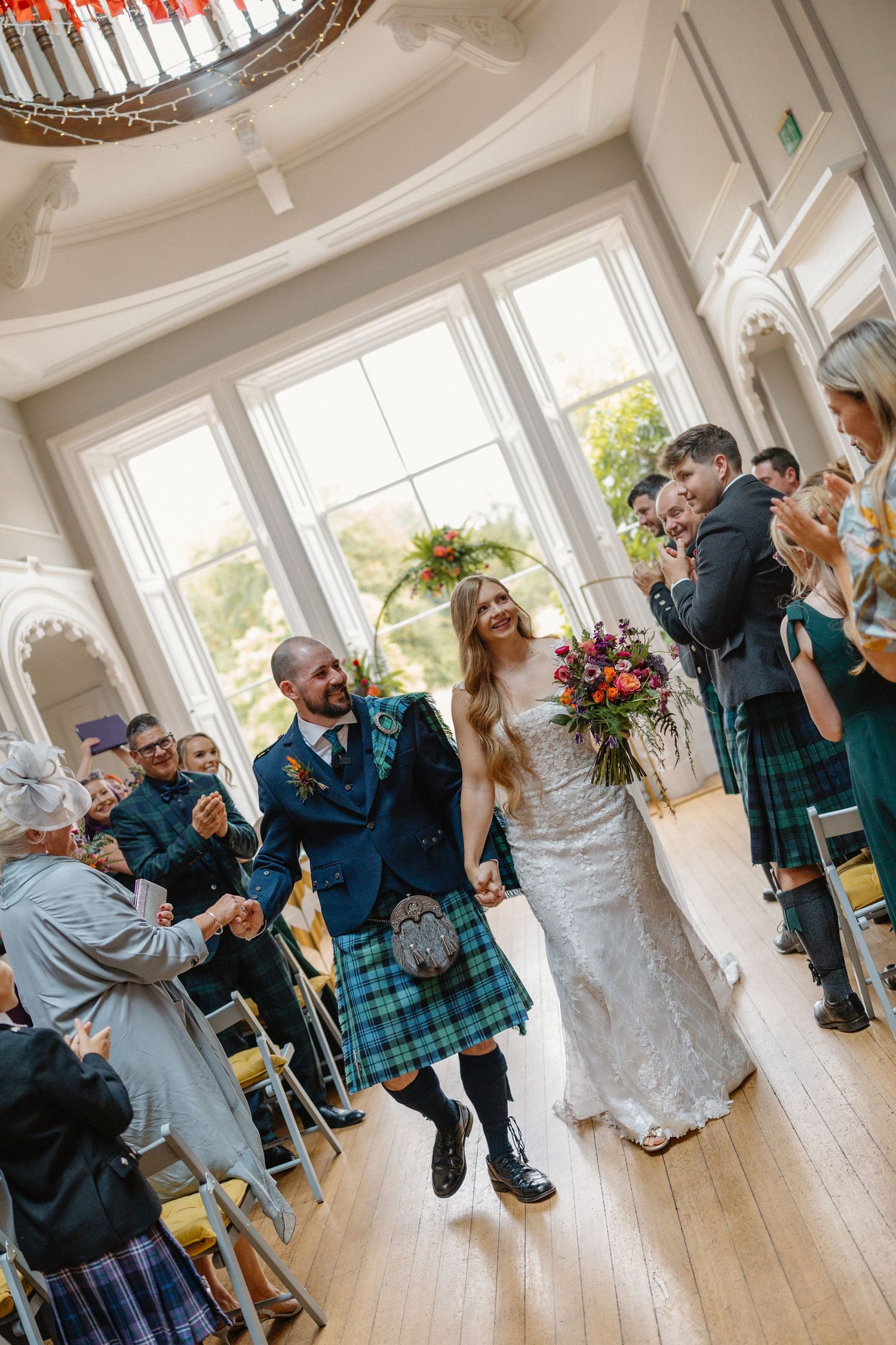 Joyful newlyweds walking down the aisle, groom in blue tartan kilt, bride holding colorful bouquet.