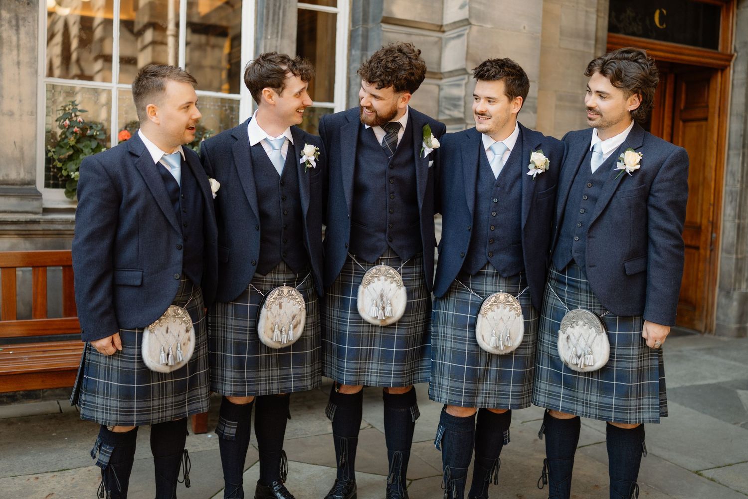 Five men in matching blue kilts and navy blazers with boutonnieres laugh together outside a stone building.