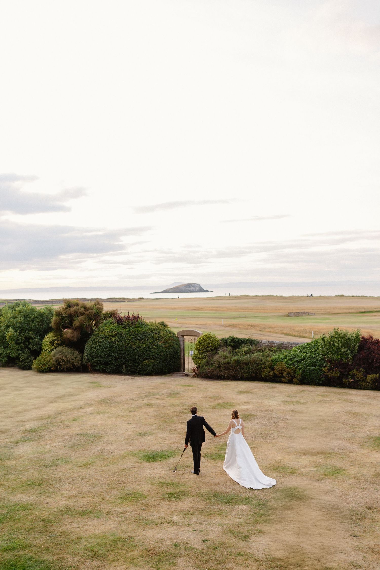 Bride and groom walking hand in hand across open grounds toward coastal landscape at sunset.