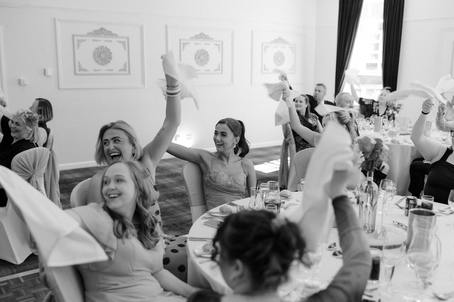 Joyful wedding guests raising hands and cheering at elegant reception dinner in black and white photo.