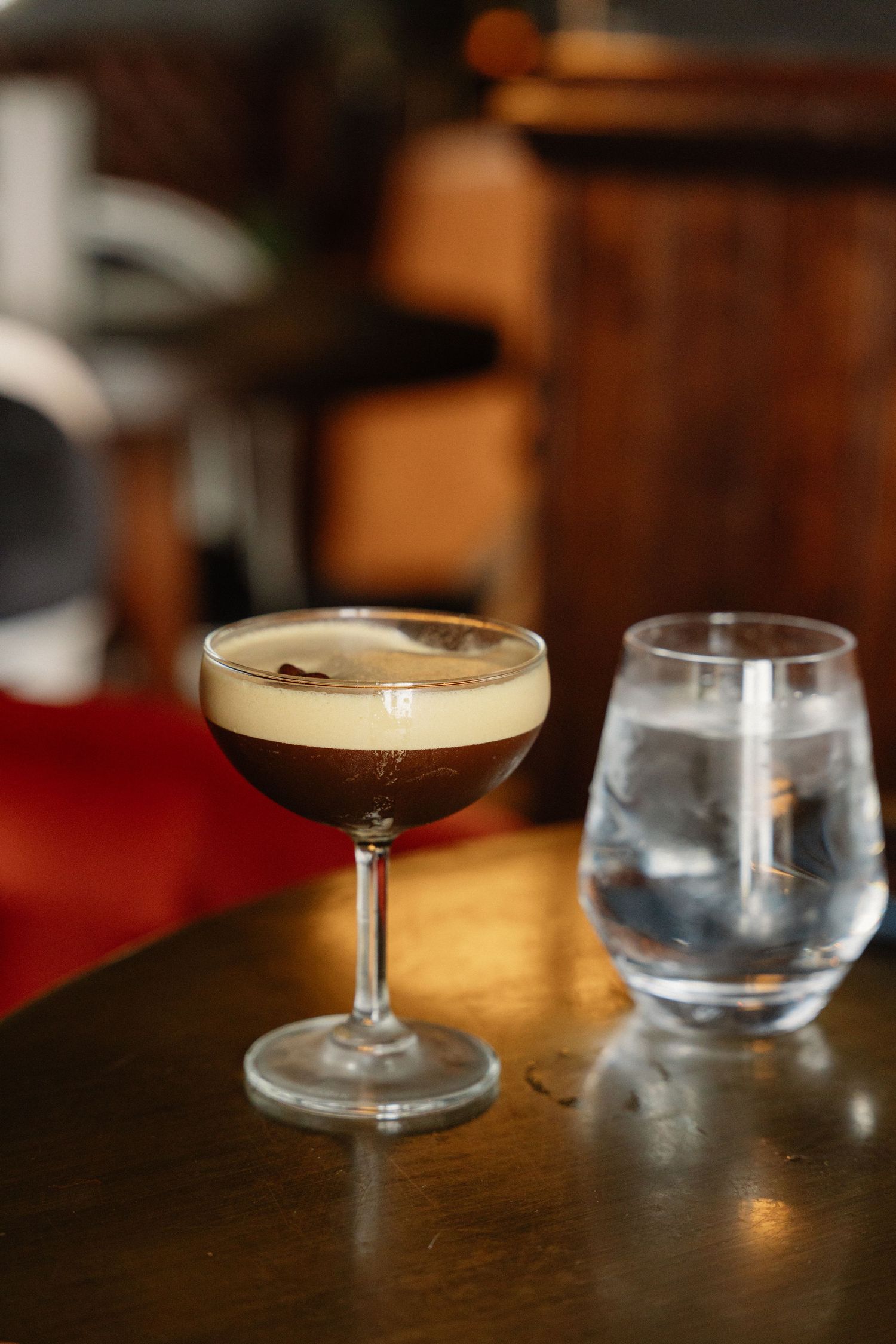 An espresso martini cocktail with creamy foam top in a coupe glass beside a water glass on a wooden bar.