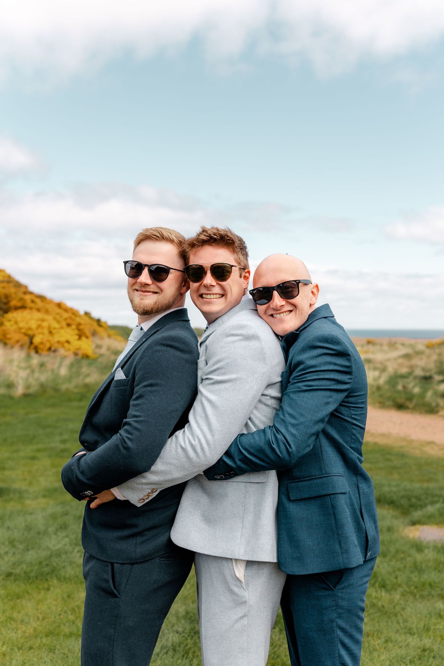 Three groomsmen in suits pose playfully together on a grassy hillside with blue sky backdrop.