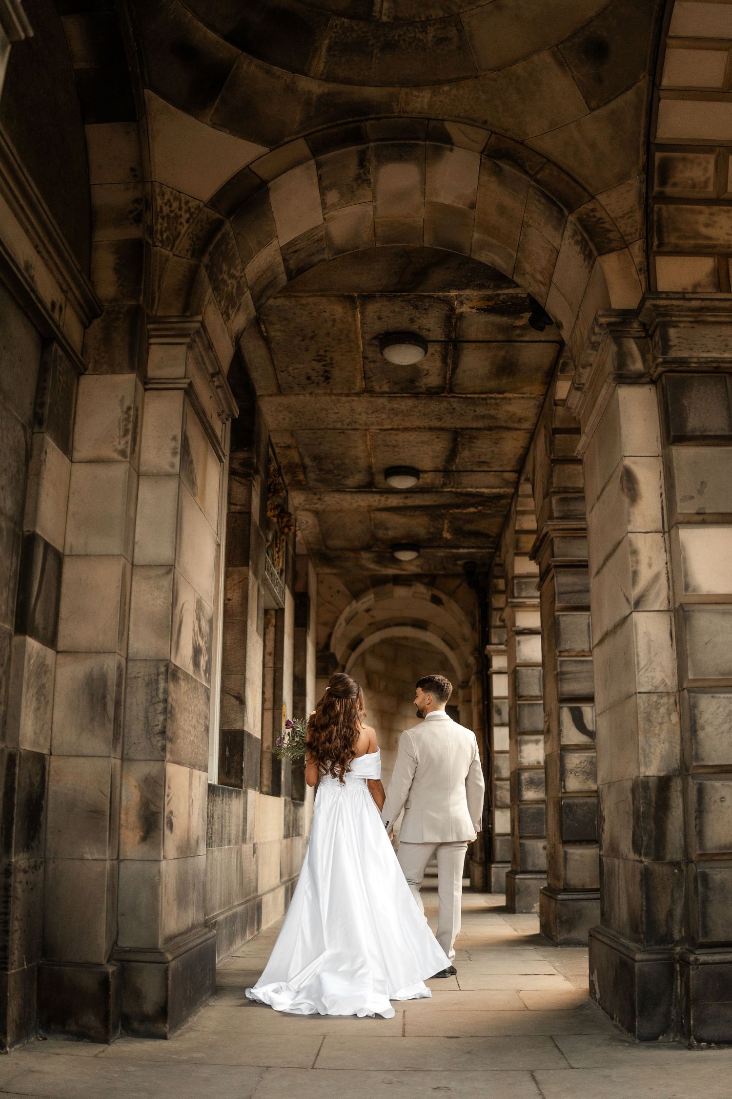 Couple walking through historic stone archway corridor in wedding attire.