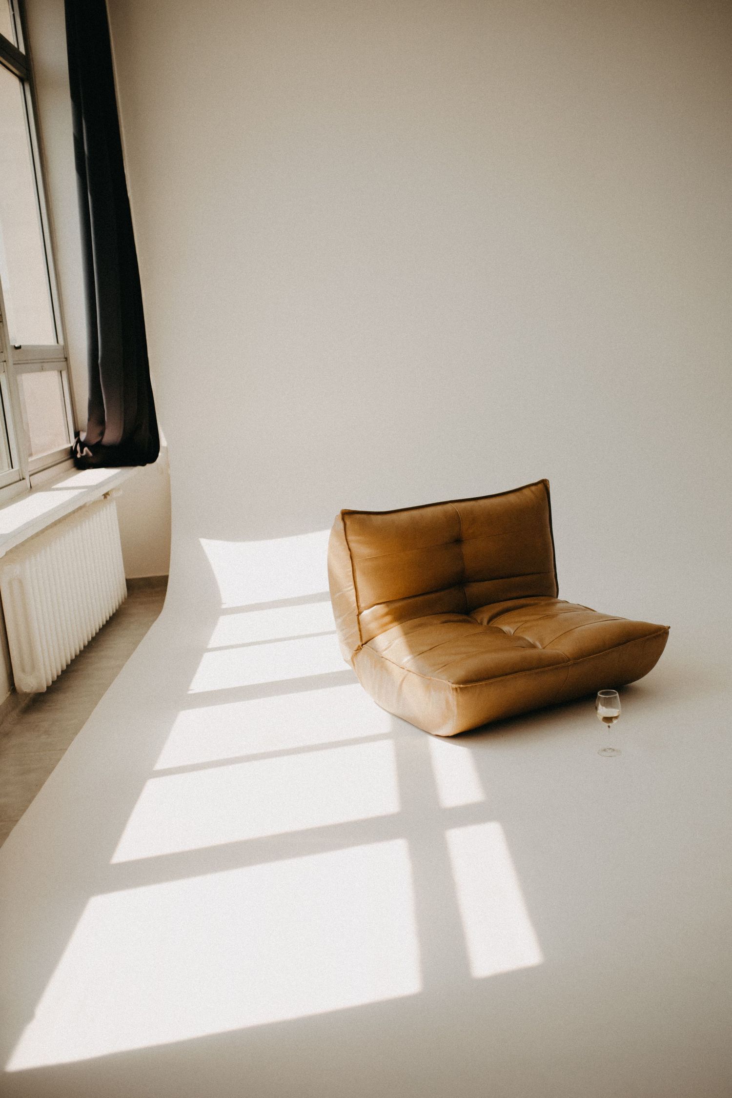 Minimalist room interior with tan leather floor cushion and dramatic window light casting shadows.