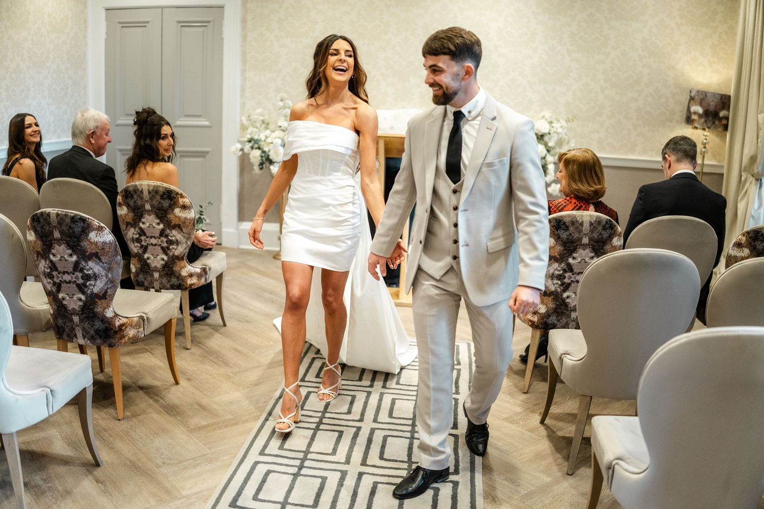 Wedding guests mingle and socialize in an elegant reception room with gray chairs and geometric floor patterns.