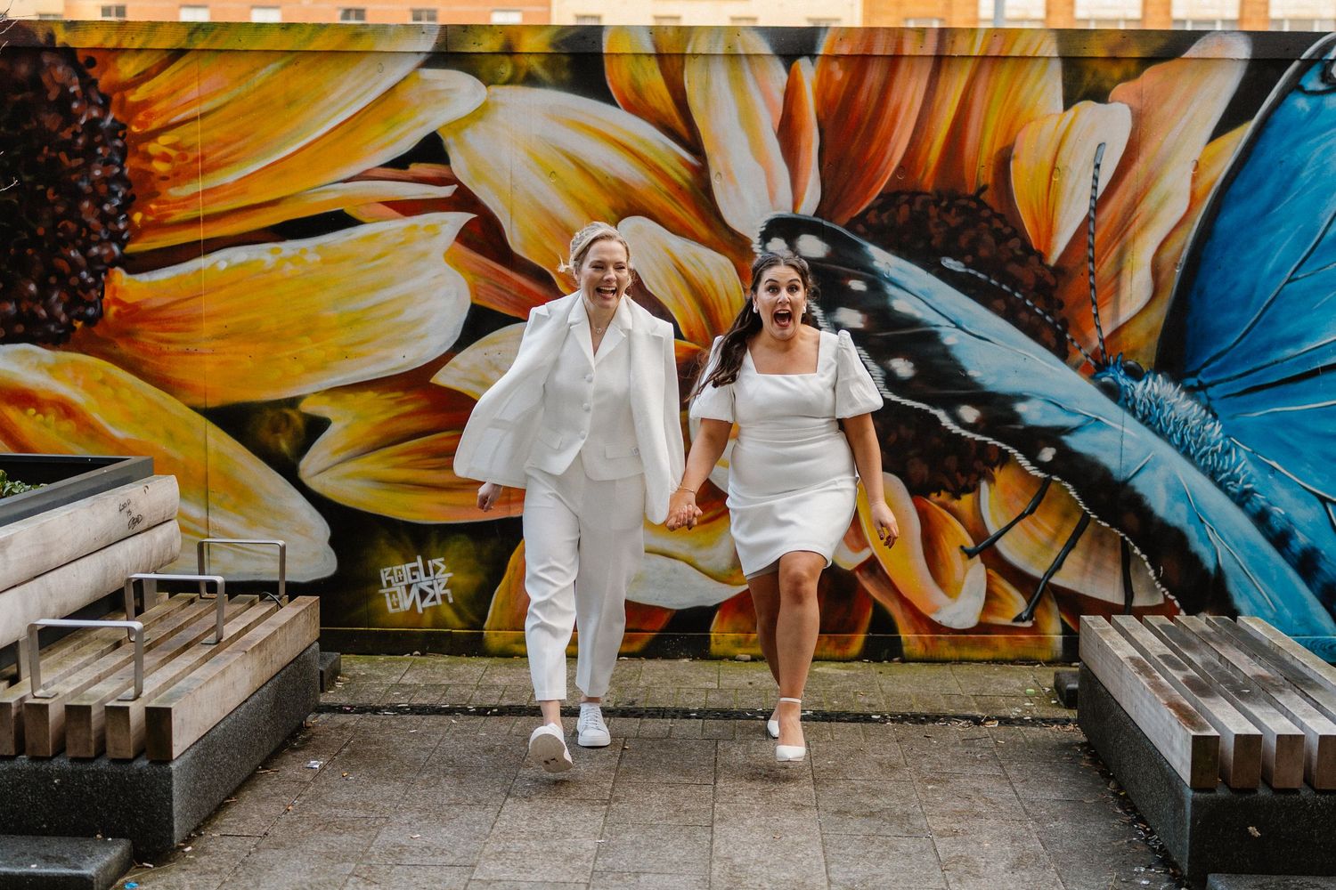 Two brides in white outfits hold hands and walk joyfully in front of a vibrant floral and butterfly mural.