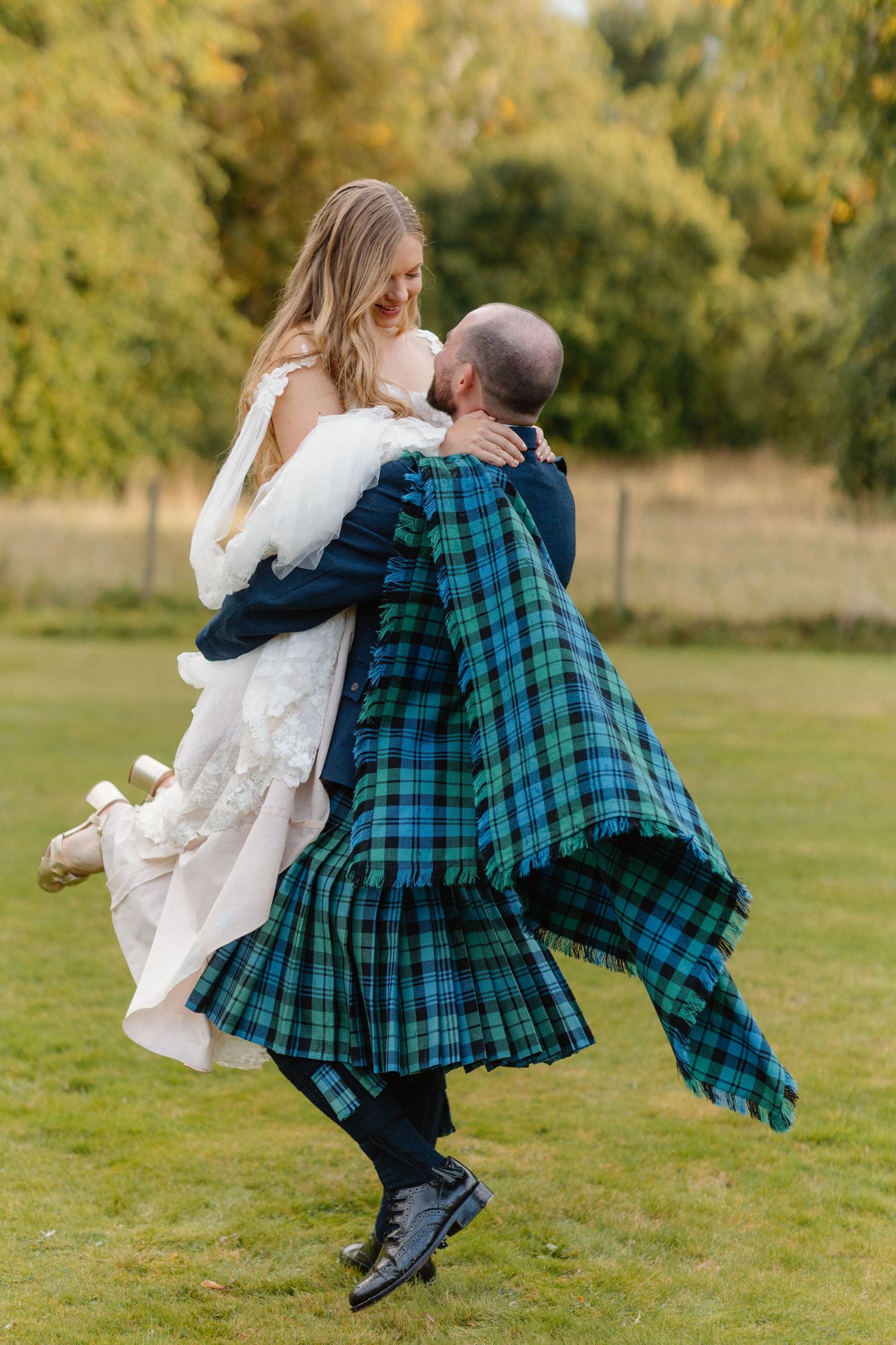 Groom in green tartan kilt joyfully lifts bride in white lace gown outdoors on lush green lawn.