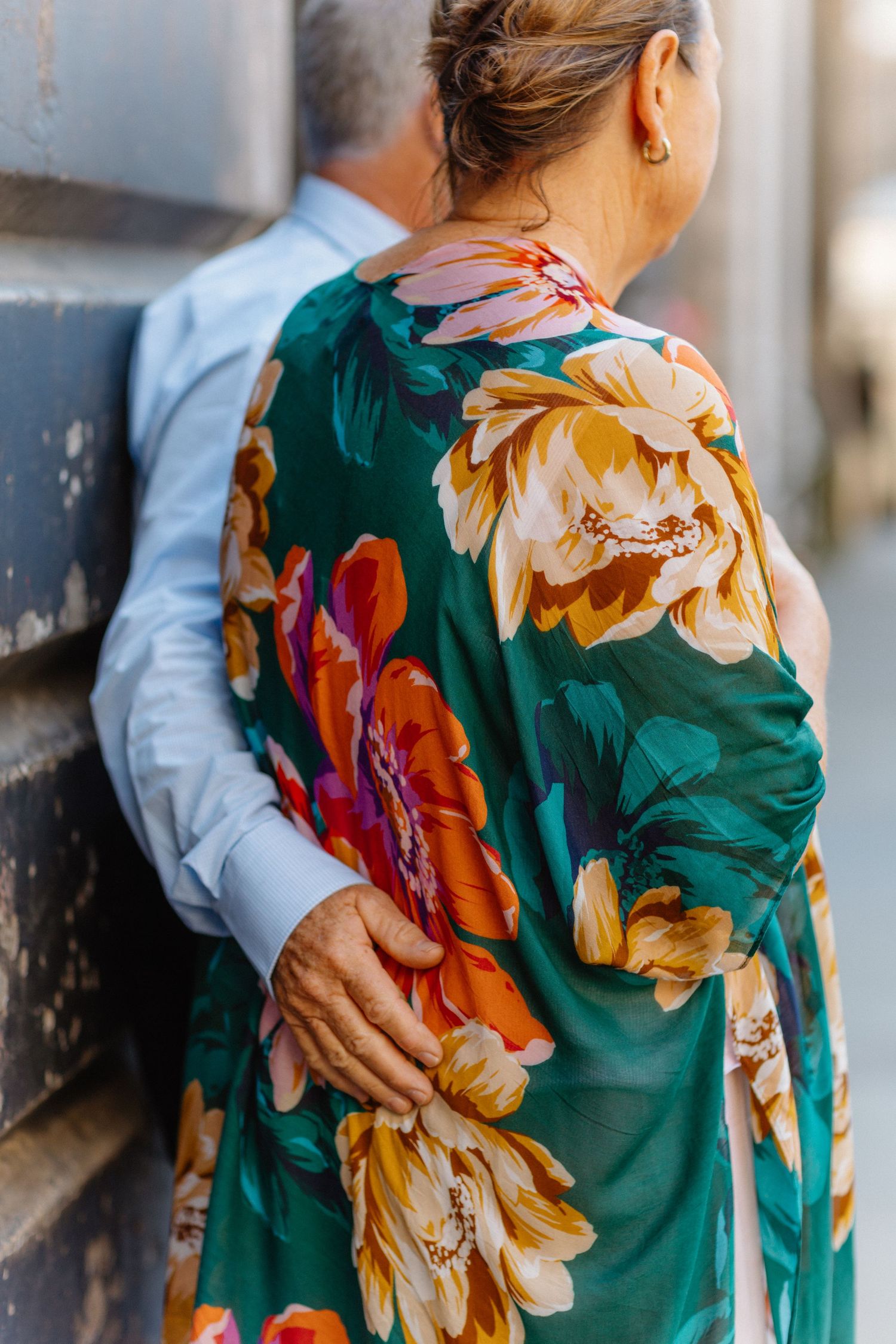 Older couple embracing outdoors, woman wearing a vibrant green floral dress with bold orange and pink blooms.