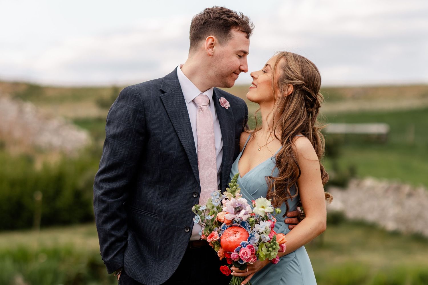 A couple in wedding attire share a romantic moment outdoors holding a colorful bouquet.