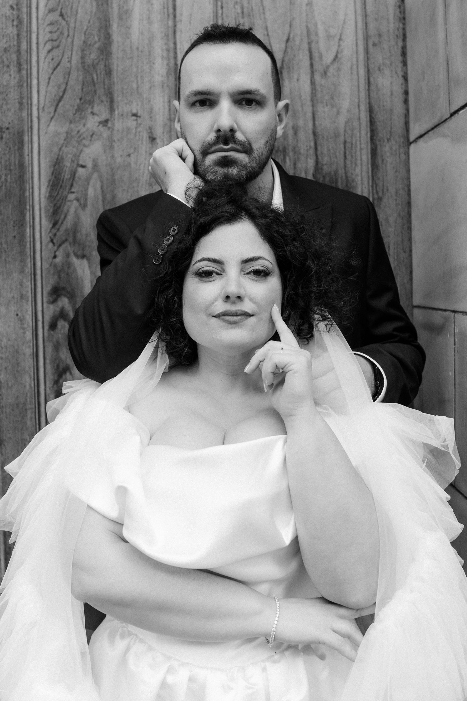 Black and white portrait of bride and groom posing elegantly against a wooden backdrop.
