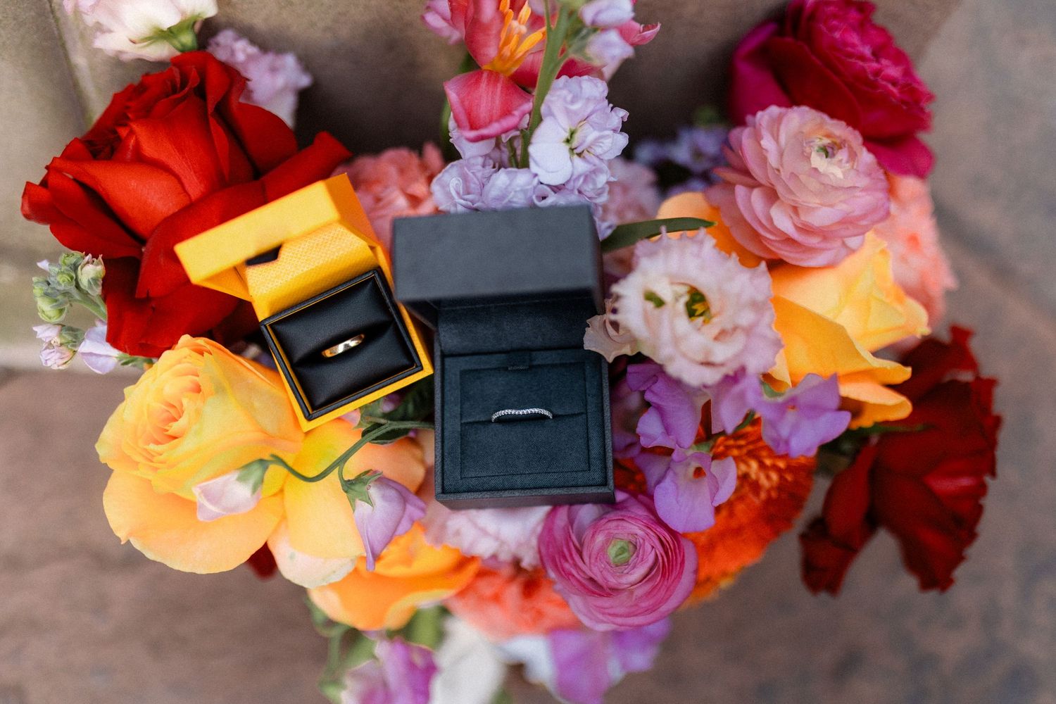 Wedding rings in yellow and black velvet boxes displayed on colorful bridal bouquet.