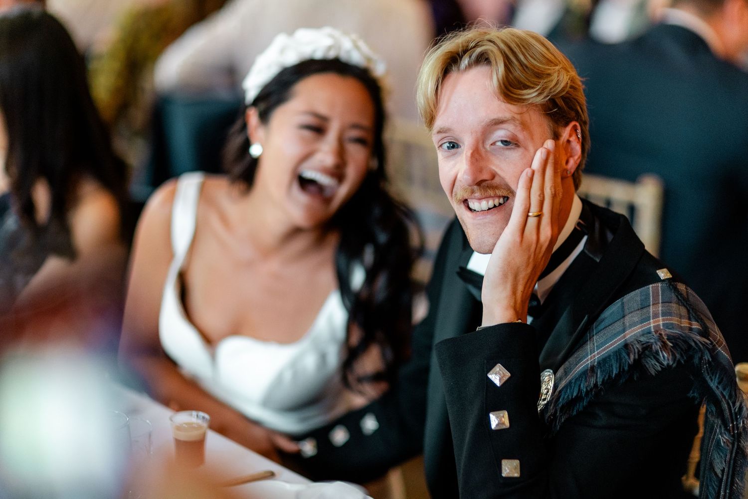 Laughing bride and grinning groom in tartan kilt sharing a joyful moment during wedding speeches.