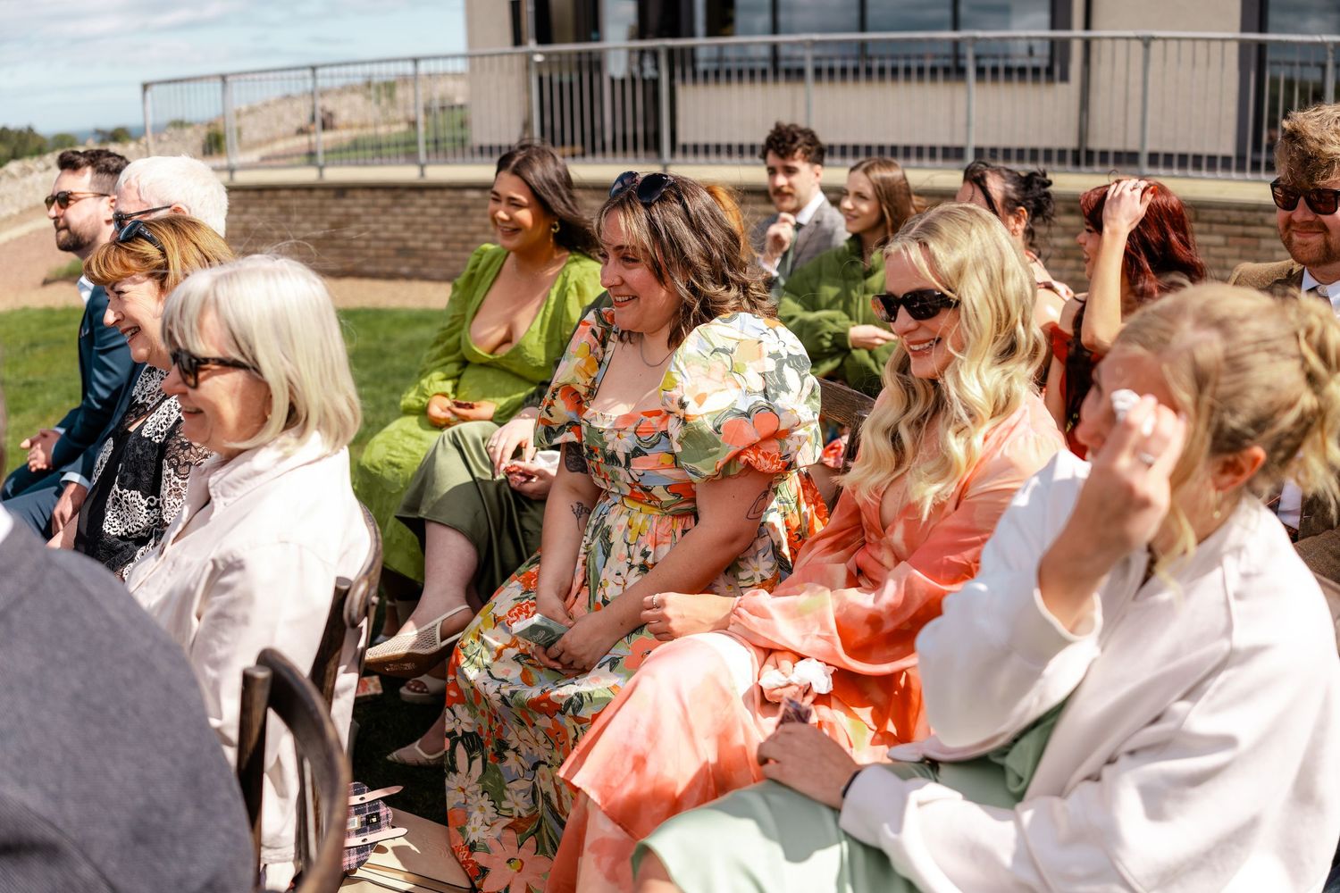 Colorfully dressed wedding guests seated outdoors enjoying a sunny wedding ceremony celebration.