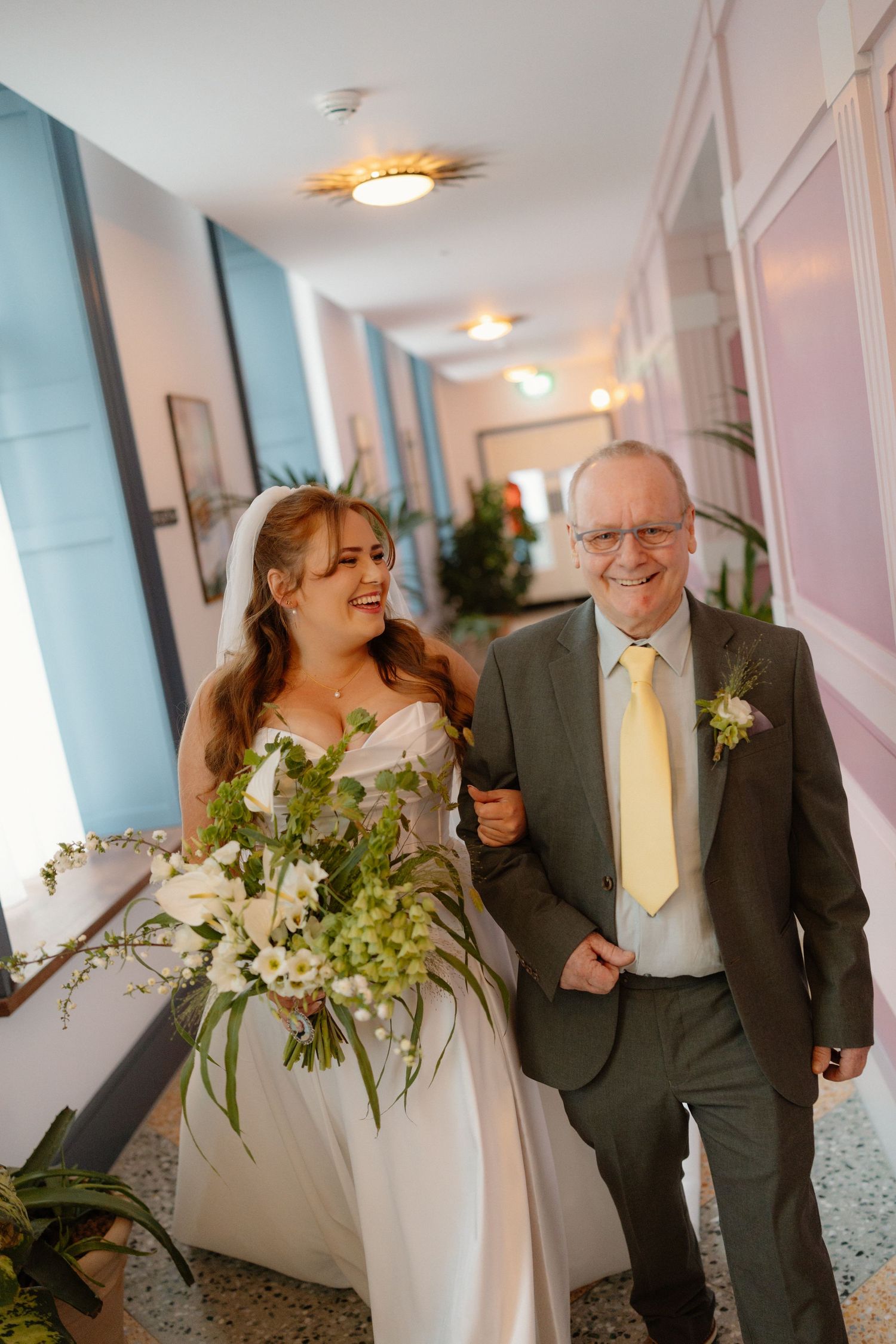 A happy bride in a white gown holding a green bouquet walks arm-in-arm with an older man in a gray suit.