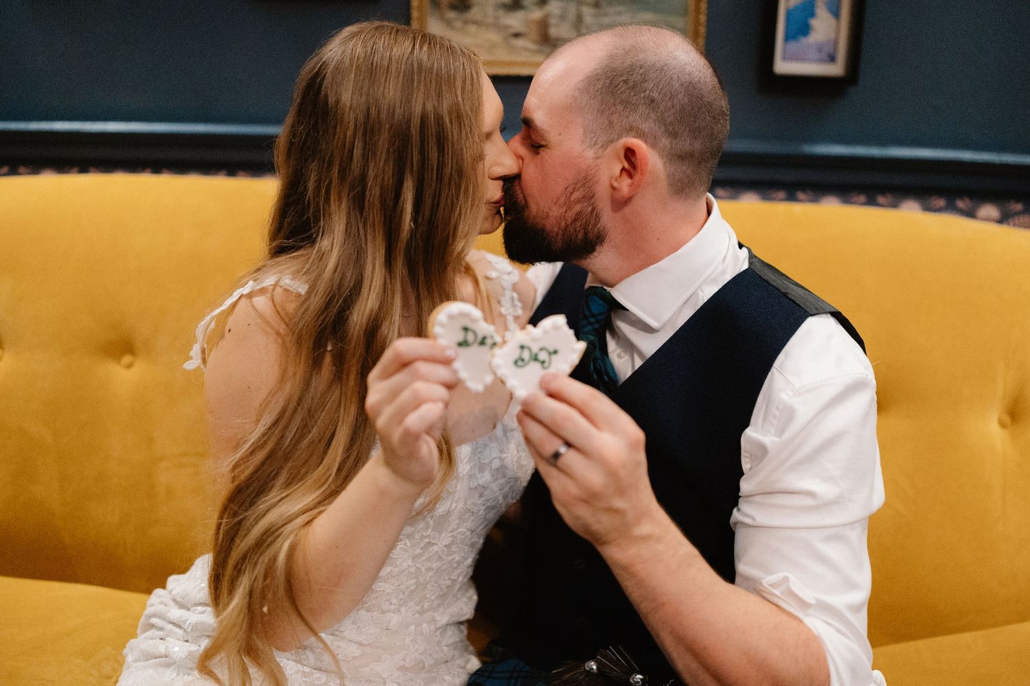 Bride and groom share a kiss on yellow sofa while holding decorative paper hearts at their wedding reception.