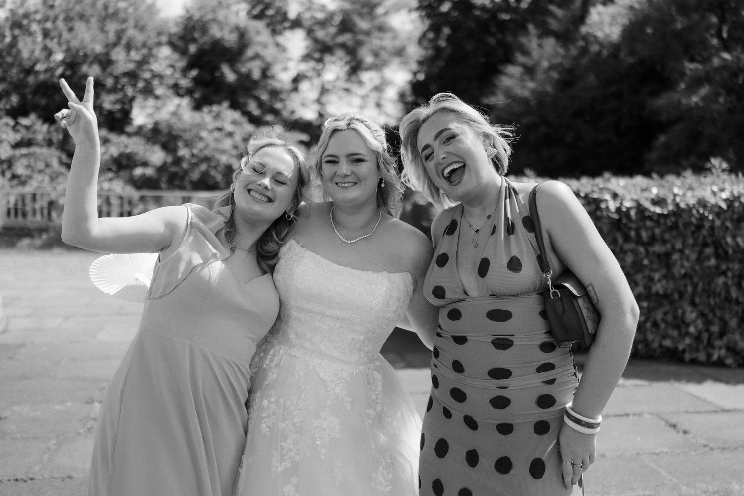 Three women pose together outdoors in black and white photo, middle woman in wedding dress flanked by friends.