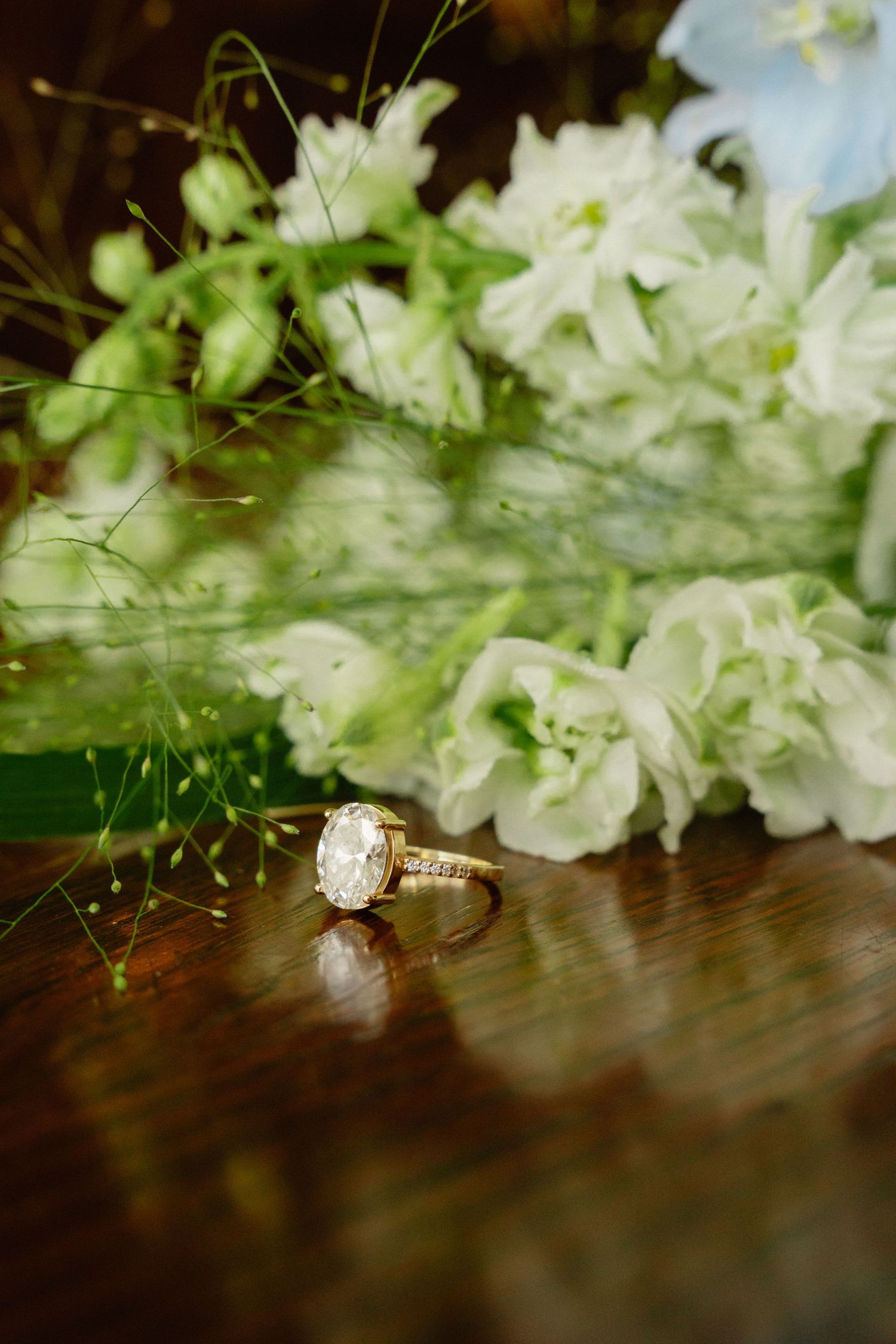 Oval diamond engagement ring in gold setting displayed on wooden surface with white flowers.