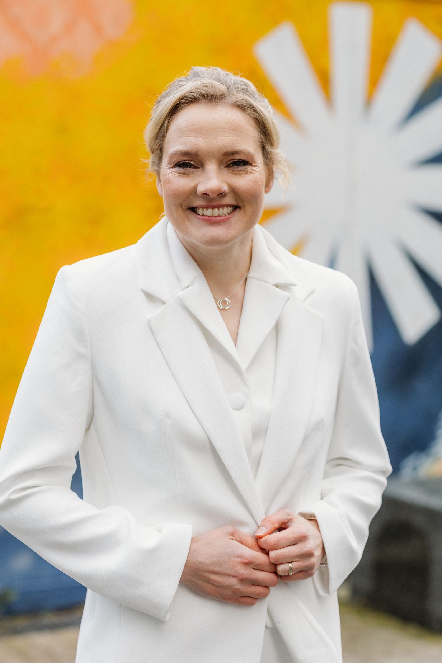 Smiling blonde bride in a white blazer poses in front of a colorful yellow street mural.