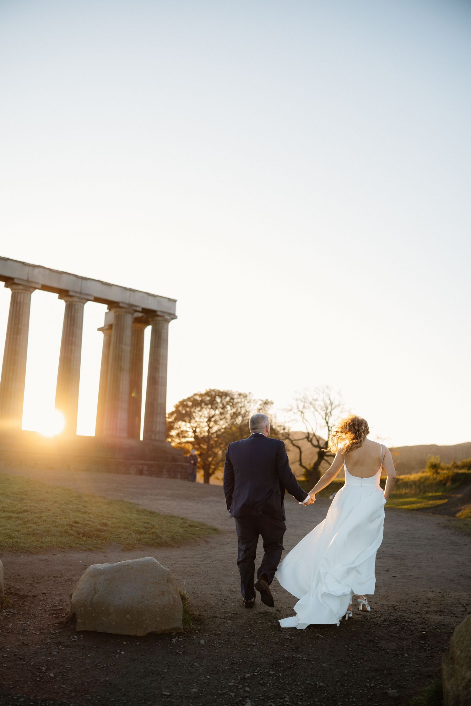 Wedding couple walking near ancient stone pillars during golden hour sunset.
