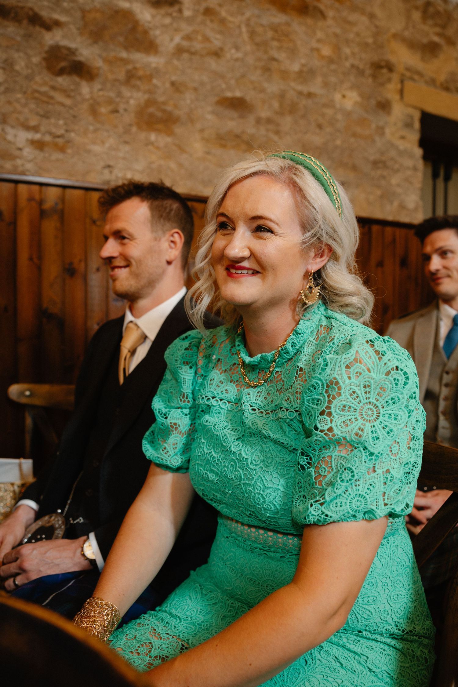 Wedding guests in emerald green lace dress enjoying ceremony in stone venue.