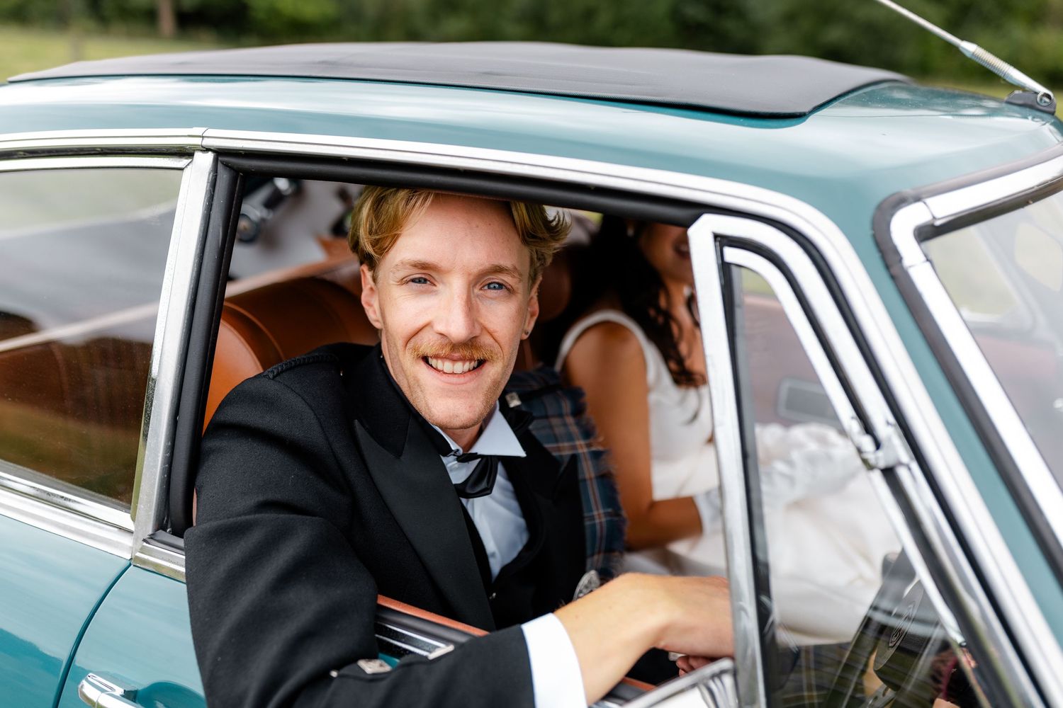A classic teal MG sports car with leather interior being driven on a country road.