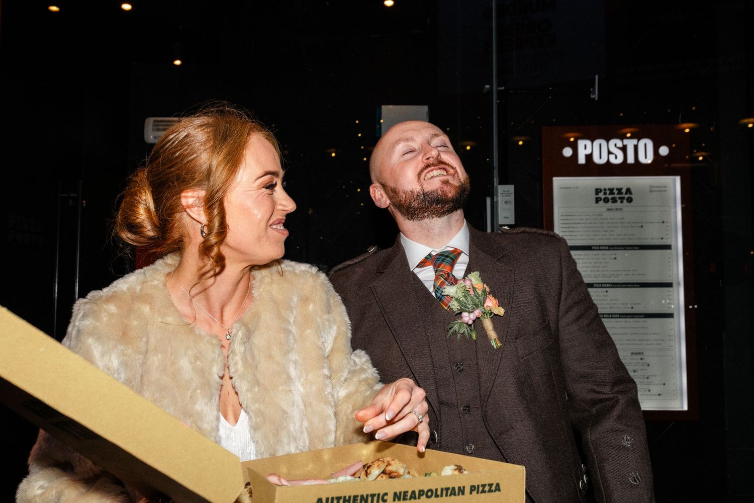 A couple laughing while sharing pizza from a cardboard box at night.