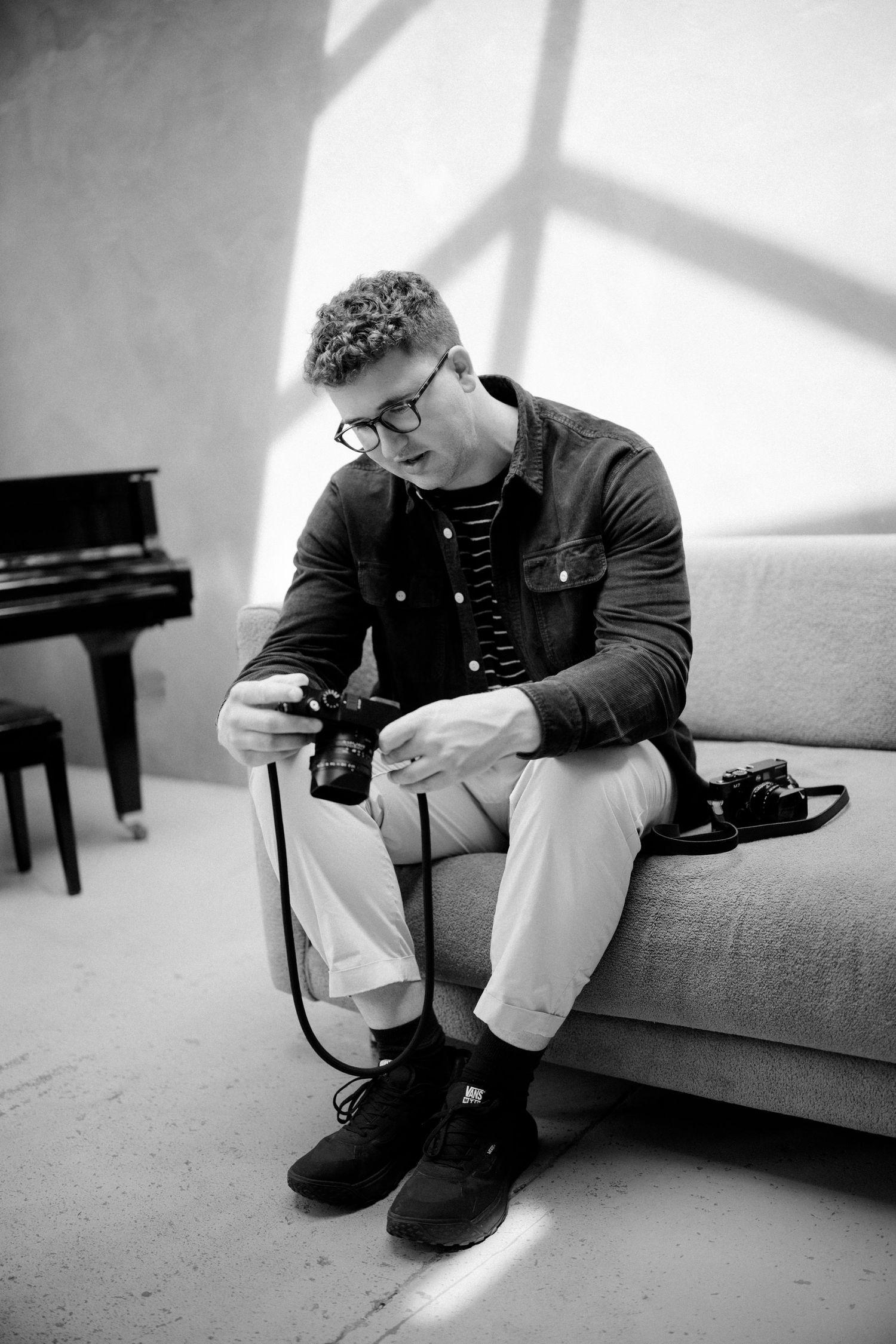 Black and white photo of person examining camera while sitting on couch near piano.