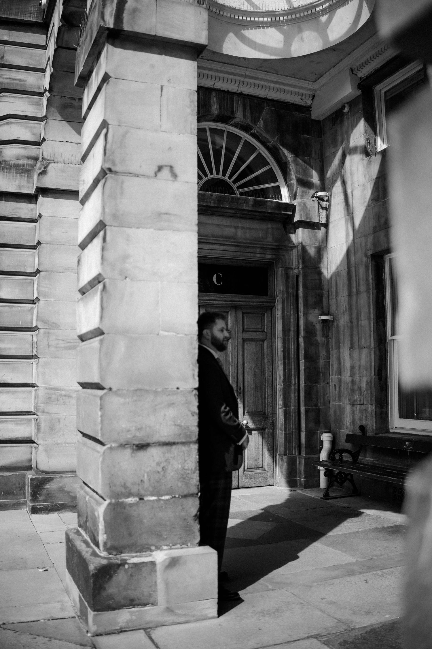 Groom in black suit stands beside ornate stone column at historic building entrance in black and white.