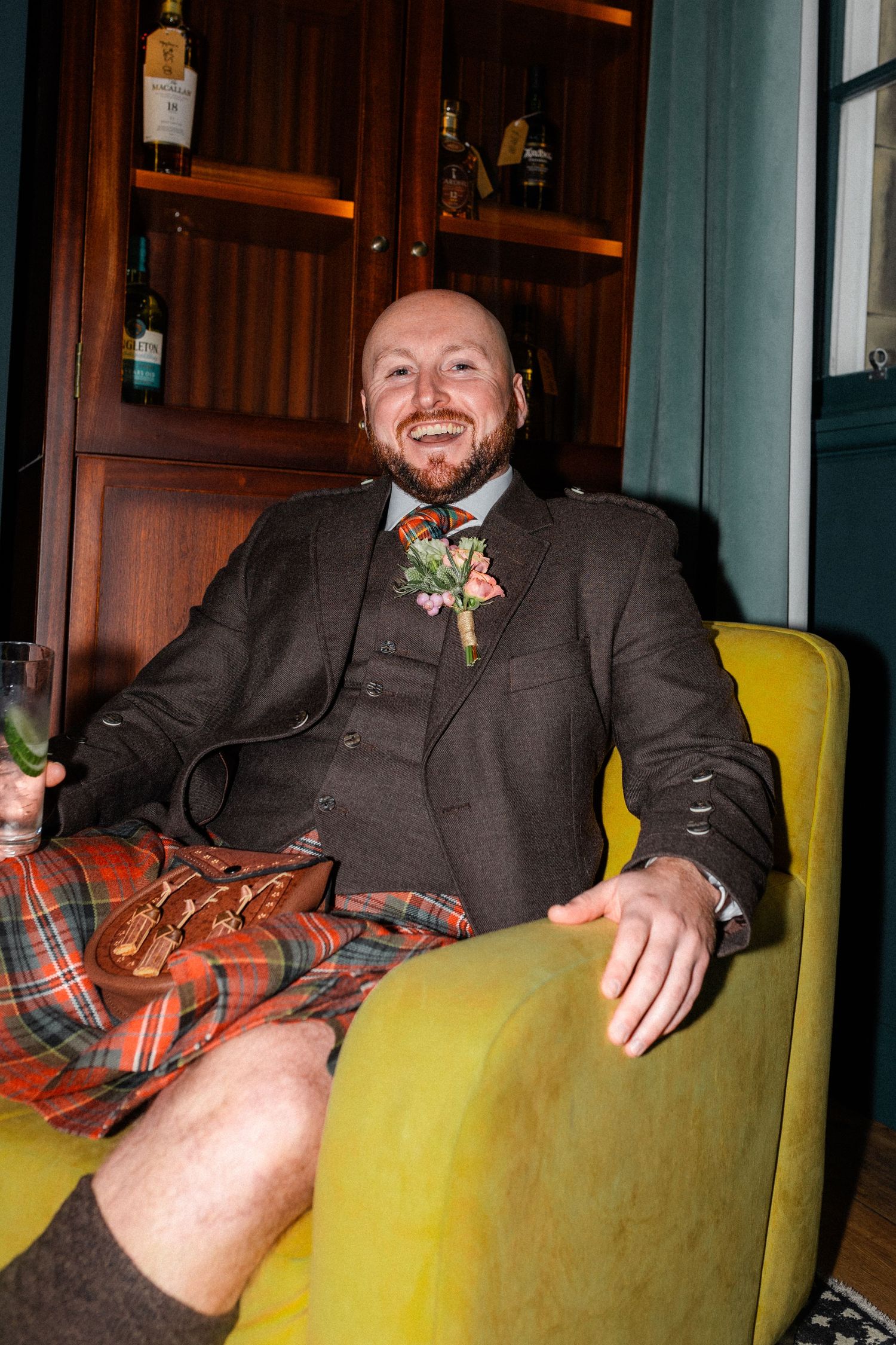 Smiling groom in tartan kilt and tweed jacket with floral boutonniere relaxes in yellow chair holding a drink.