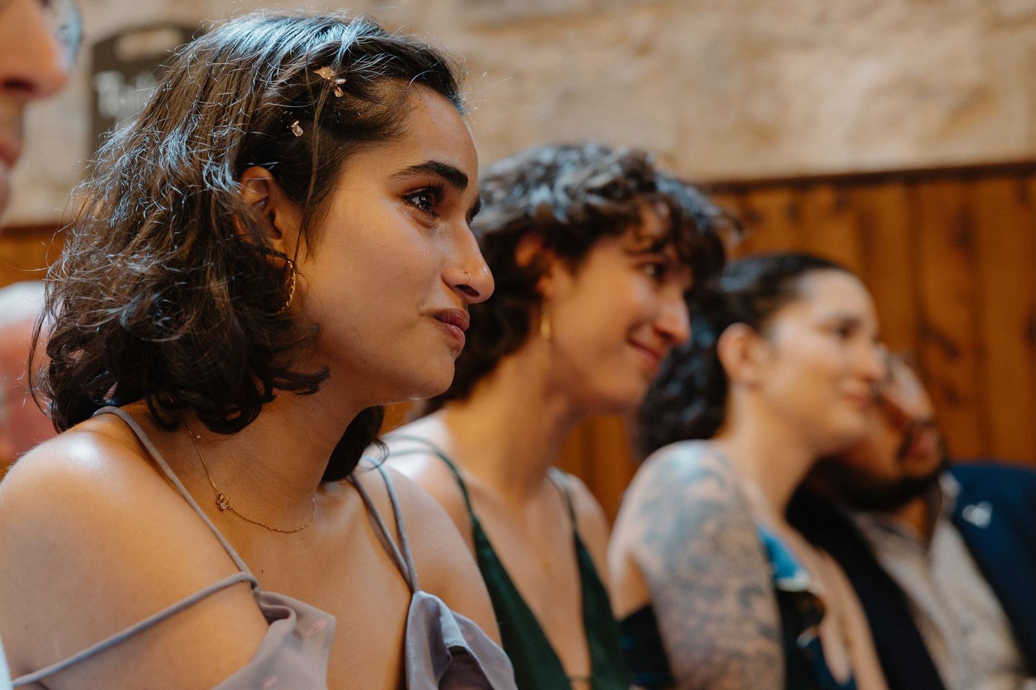 Young woman in profile view at a wedding ceremony, with guests seated behind her in a stone venue.