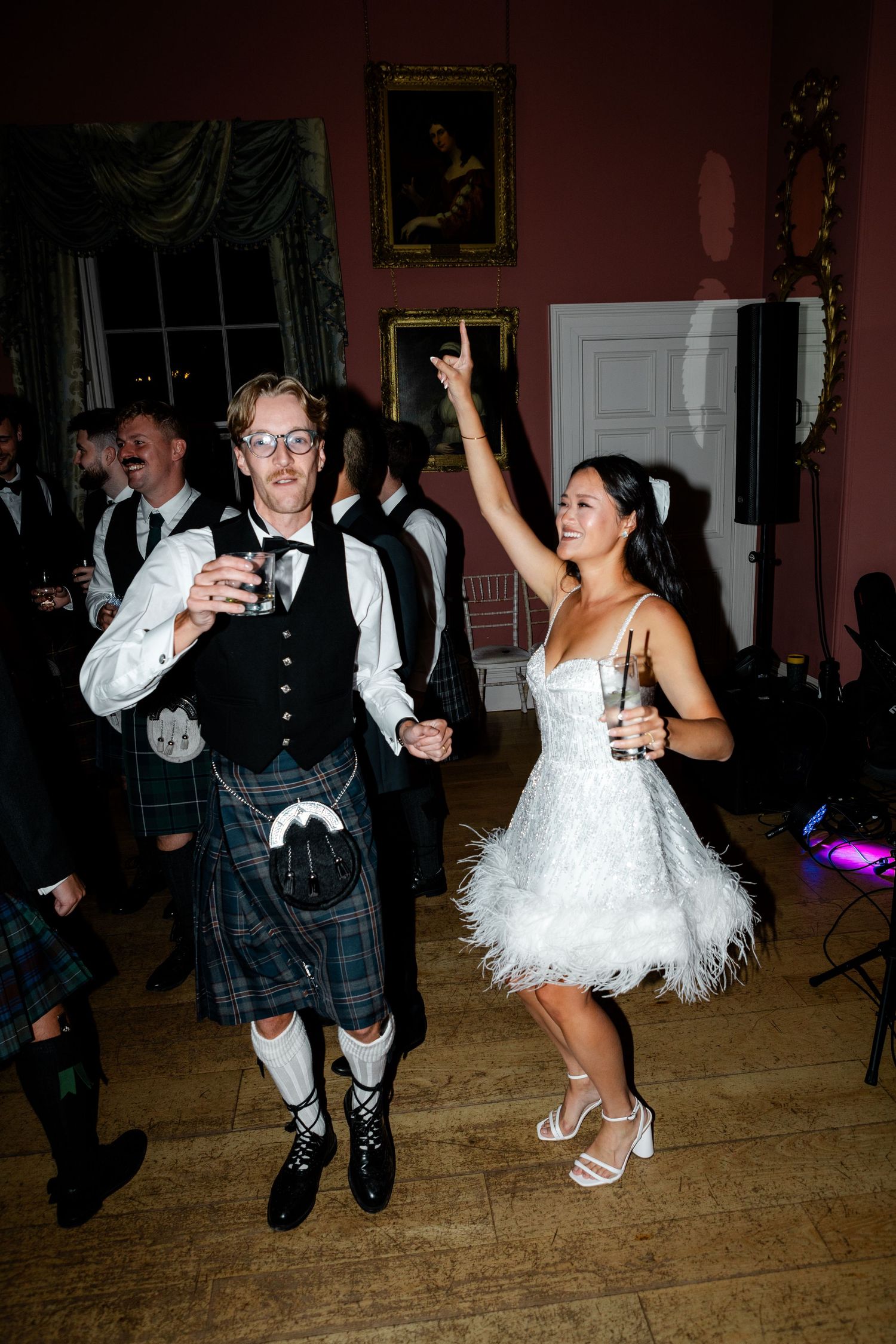 Bride in white feather mini dress dances with groom in Scottish kilt at elegant wedding reception.