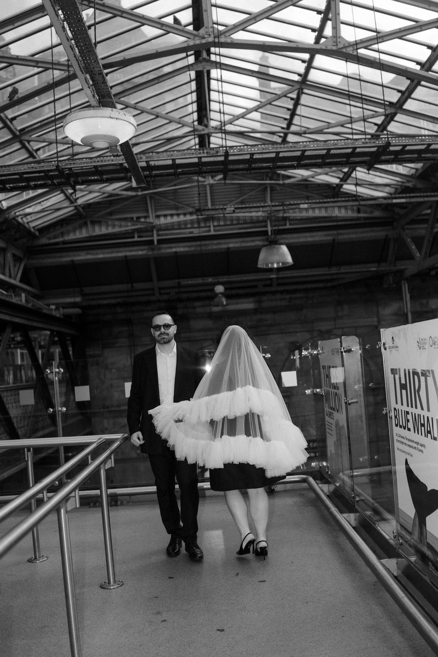 Black and white photo of a bride in short dress and veil walking with groom in suit through a glass-roofed venue.