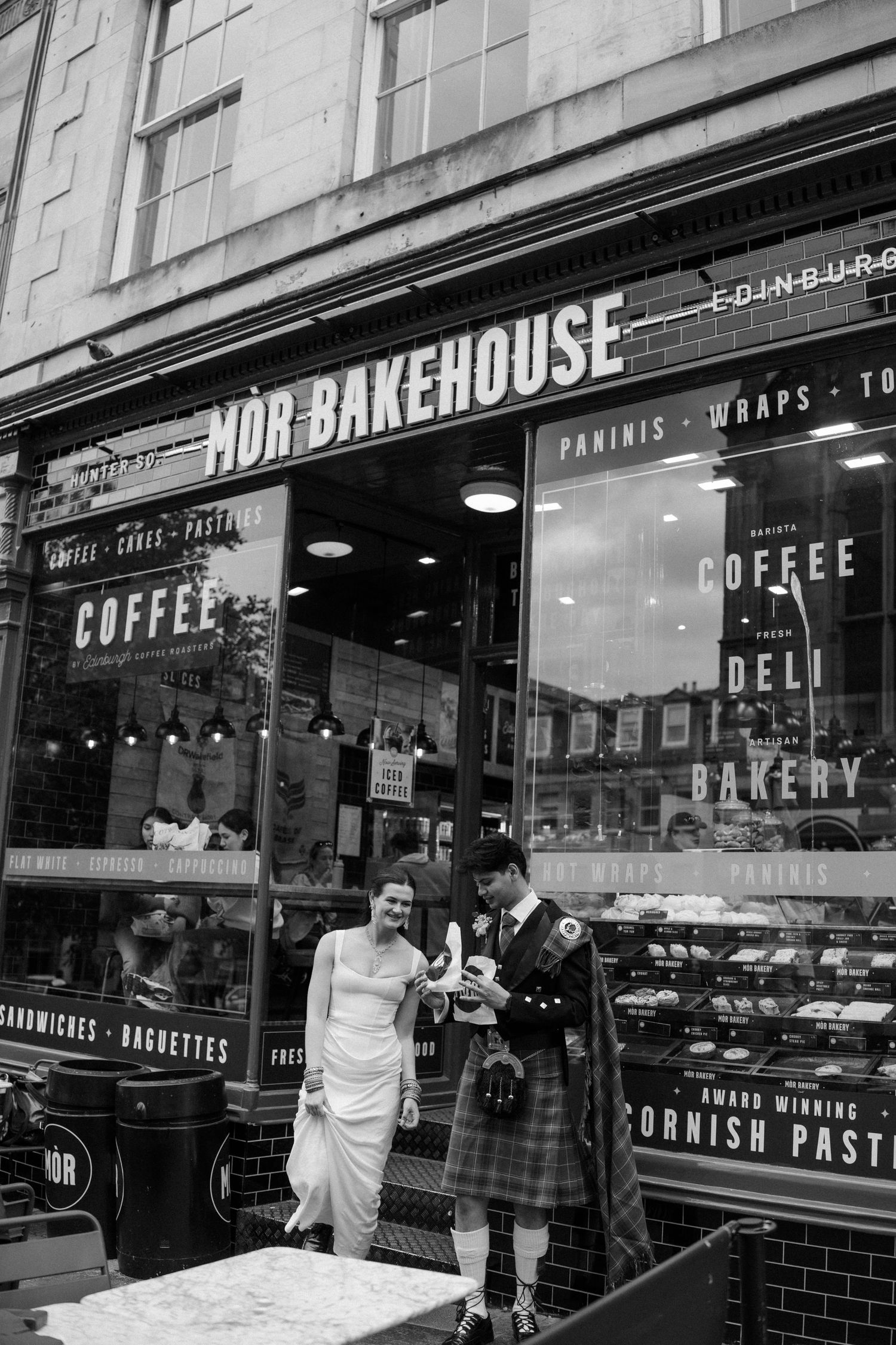Black and white photo of newlyweds in Scottish wedding attire outside Mor Bakehouse in Edinburgh.