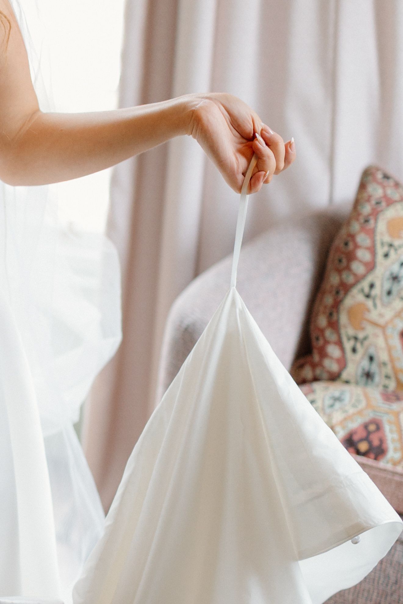 Bride holding a delicate white wedding dress by its strap near a sunlit window with soft curtains.