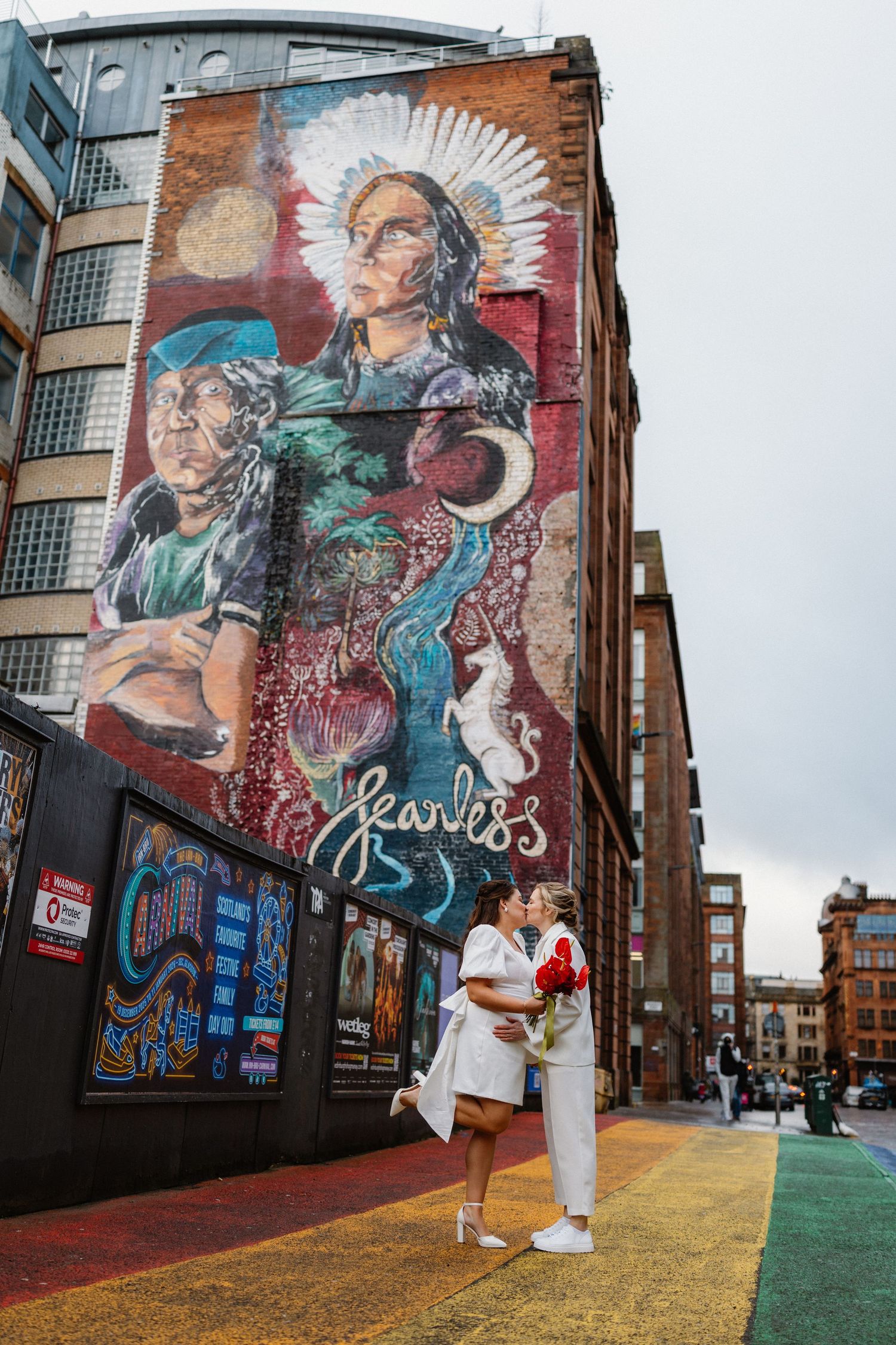 Two brides share a kiss in front of a large colorful Fearless street mural in the city.