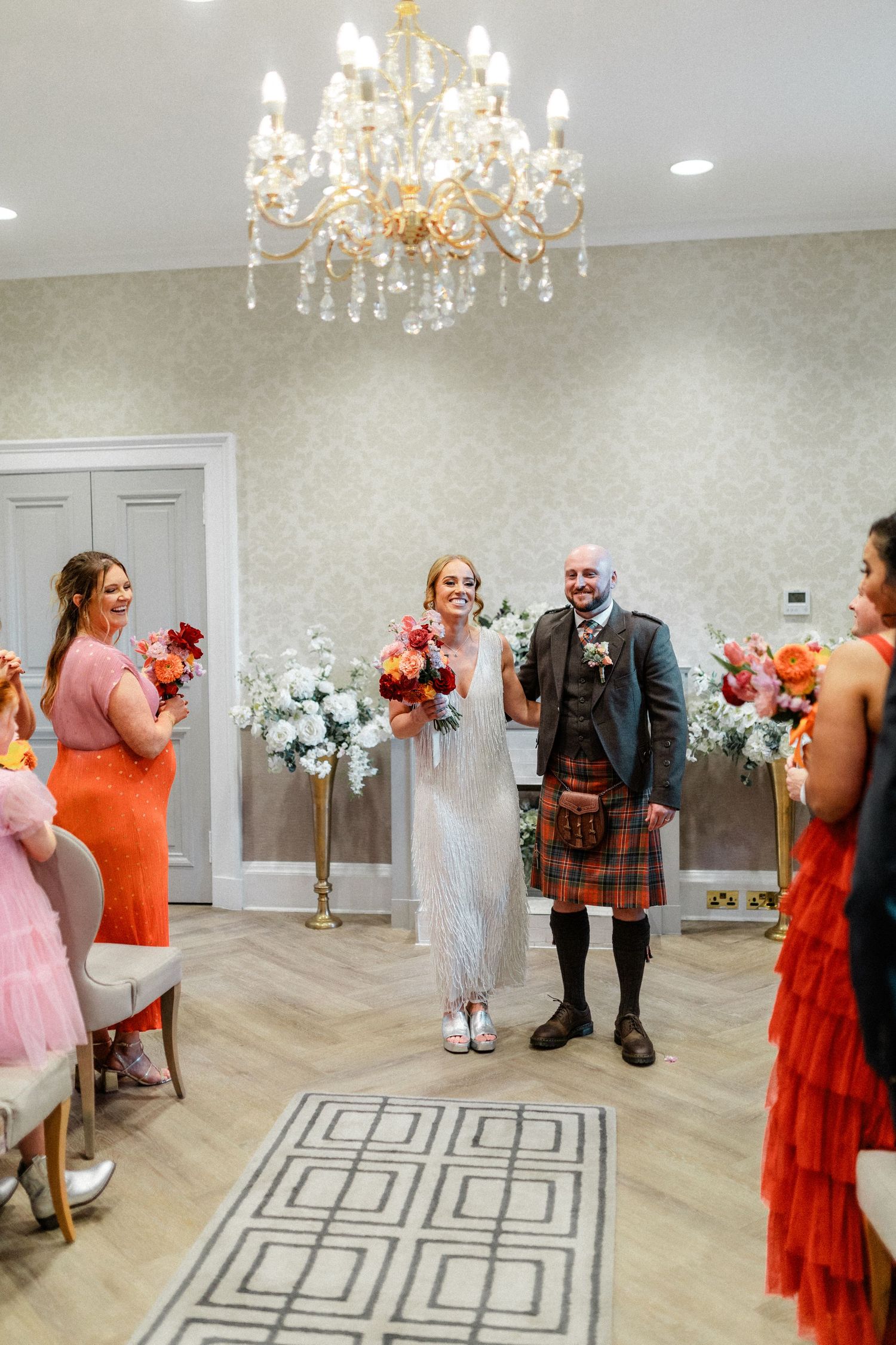 Wedding party in formal attire dancing in an elegant room with chandeliers and geometric floor pattern.