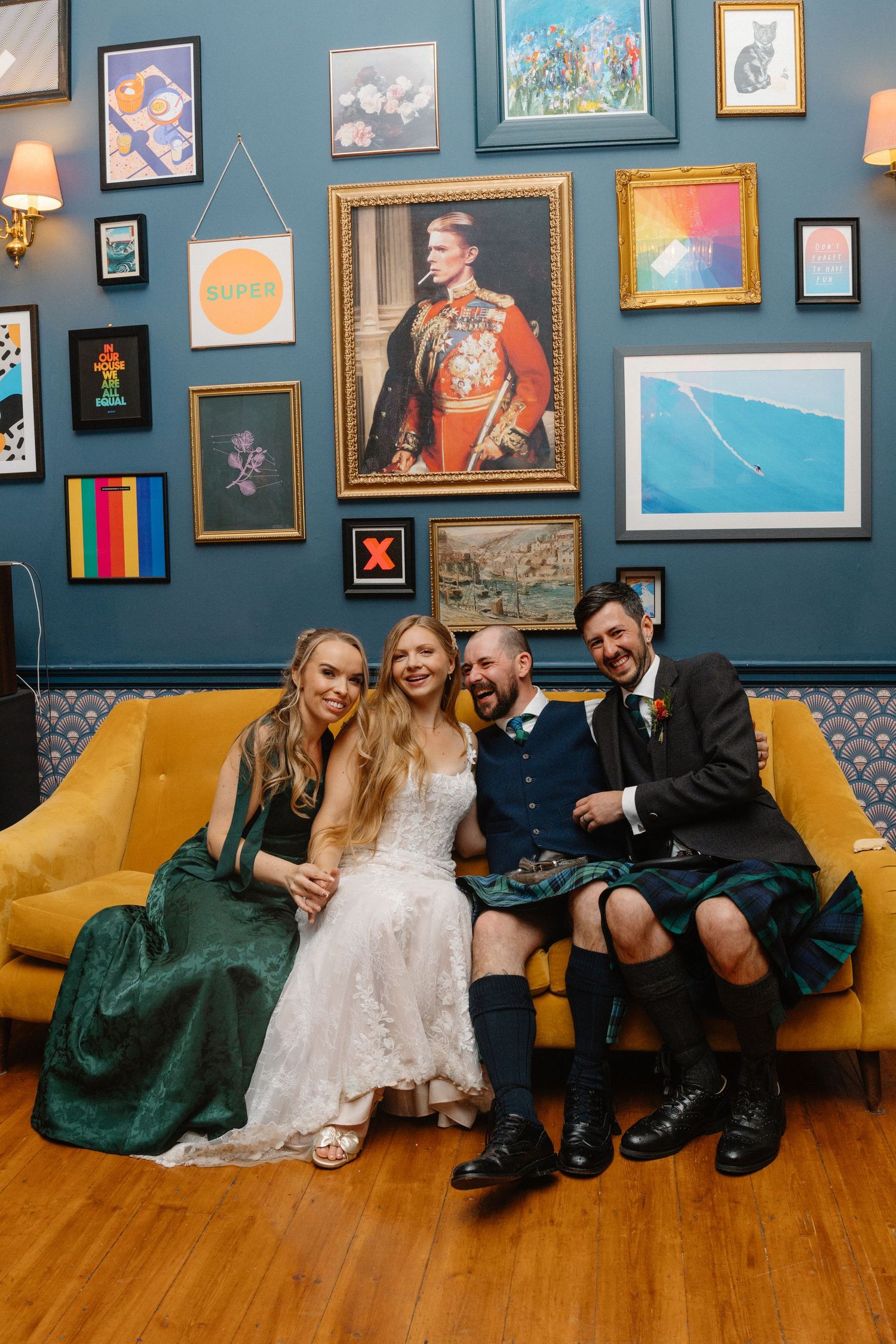 Wedding party lounging on yellow couch against blue wall with eclectic art gallery.
