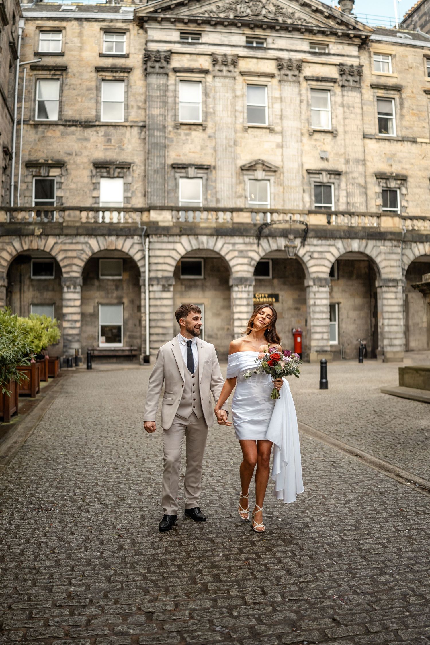 A couple walks together on cobblestone streets by a grand historic building with arched columns.