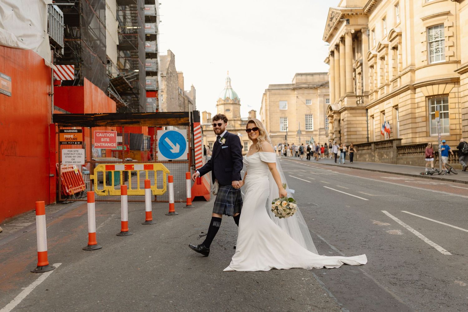 Bride and groom walk hand in hand through city street with construction barriers on their wedding day.