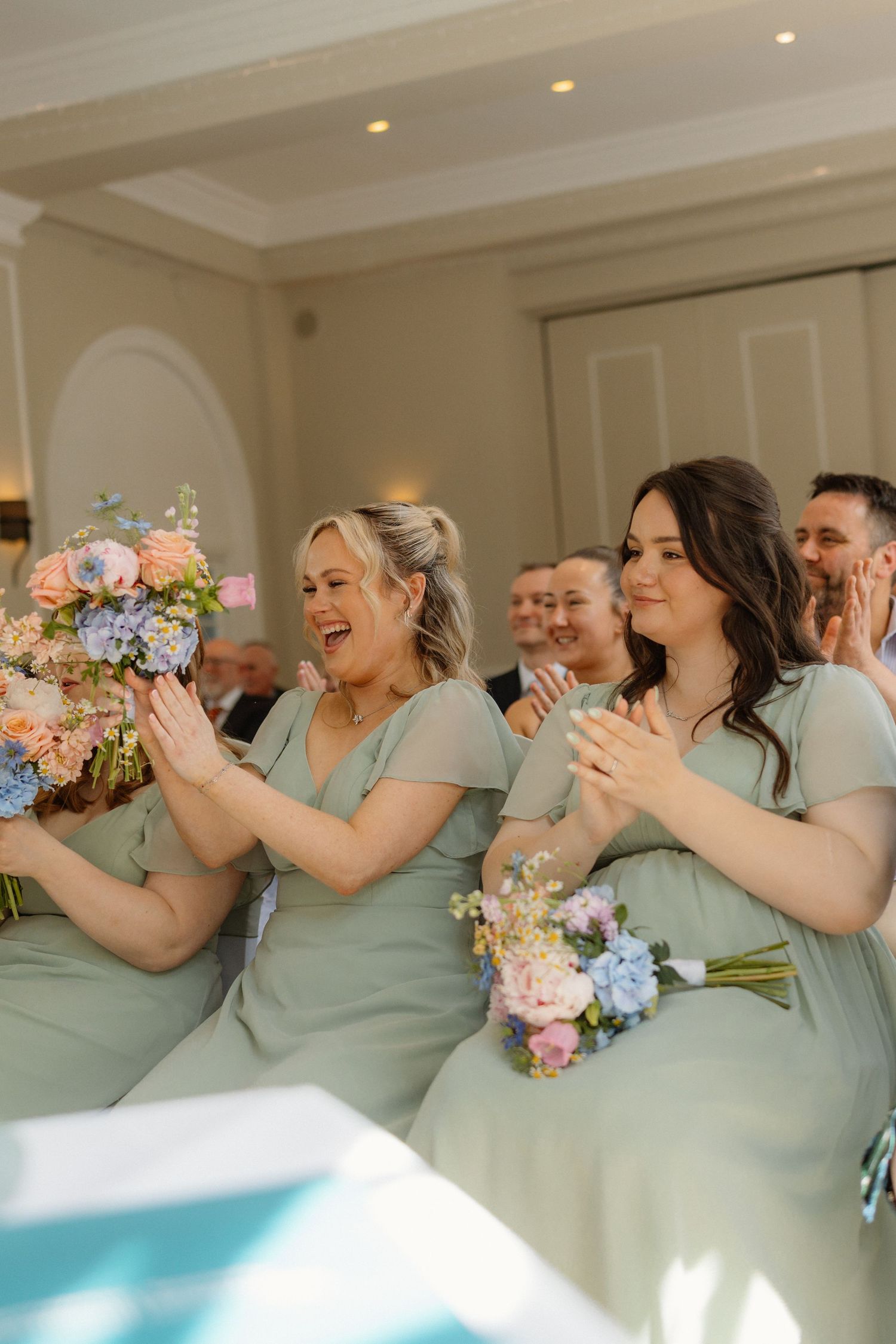 Bridesmaids in sage green dresses clapping and holding pink and blue bouquets during a wedding ceremony indoors.