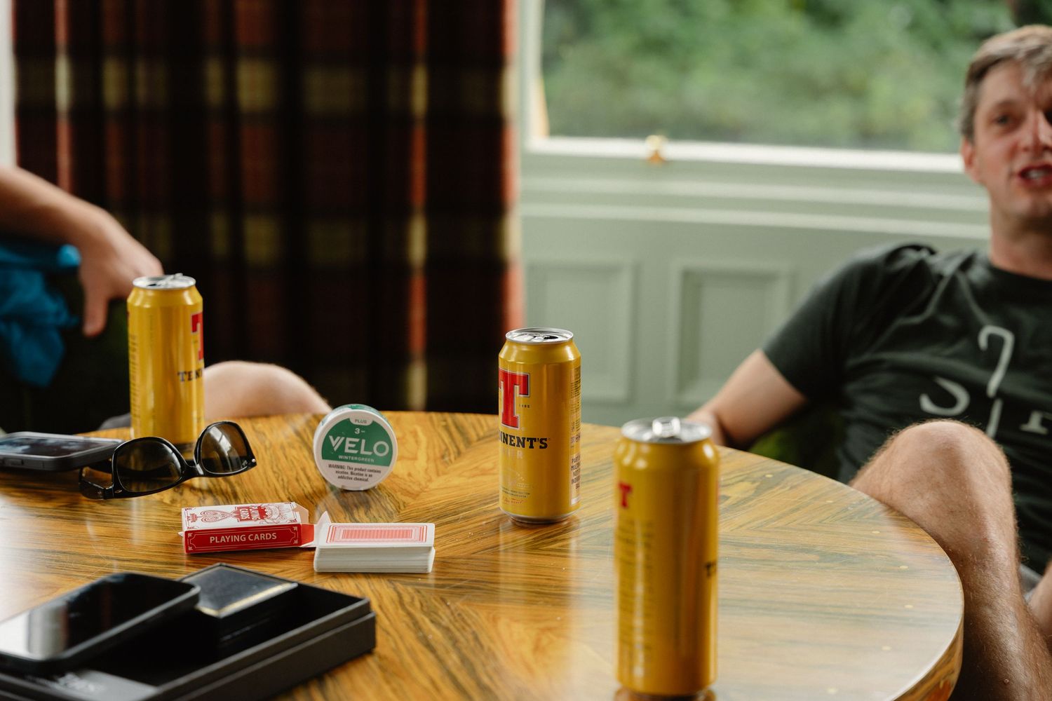 Two men sitting at a wooden pub table with beer cans and a Veld sticker, relaxing indoors near a window.