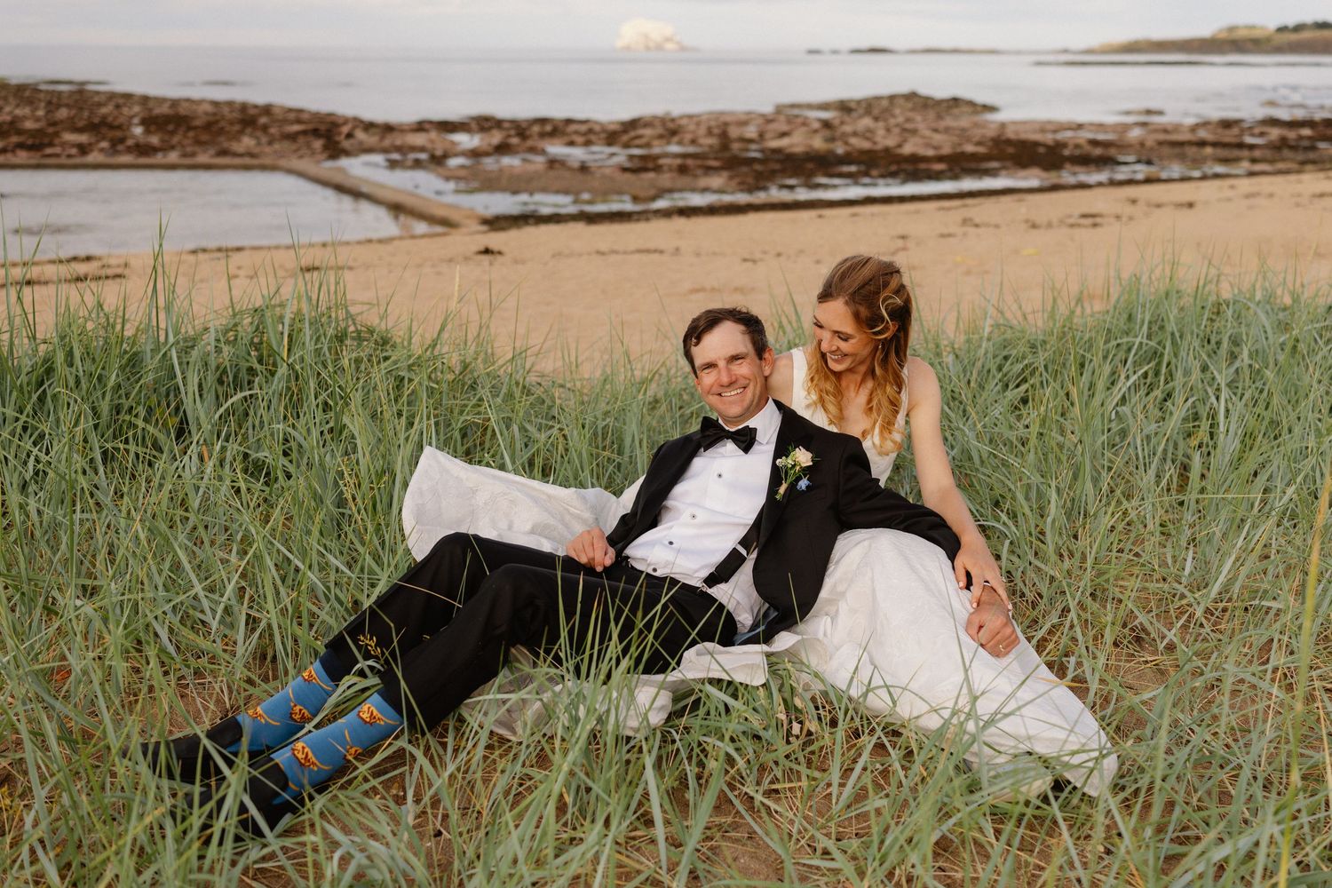Bride and groom sitting together in coastal grass dunes by the beach at sunset in wedding attire.