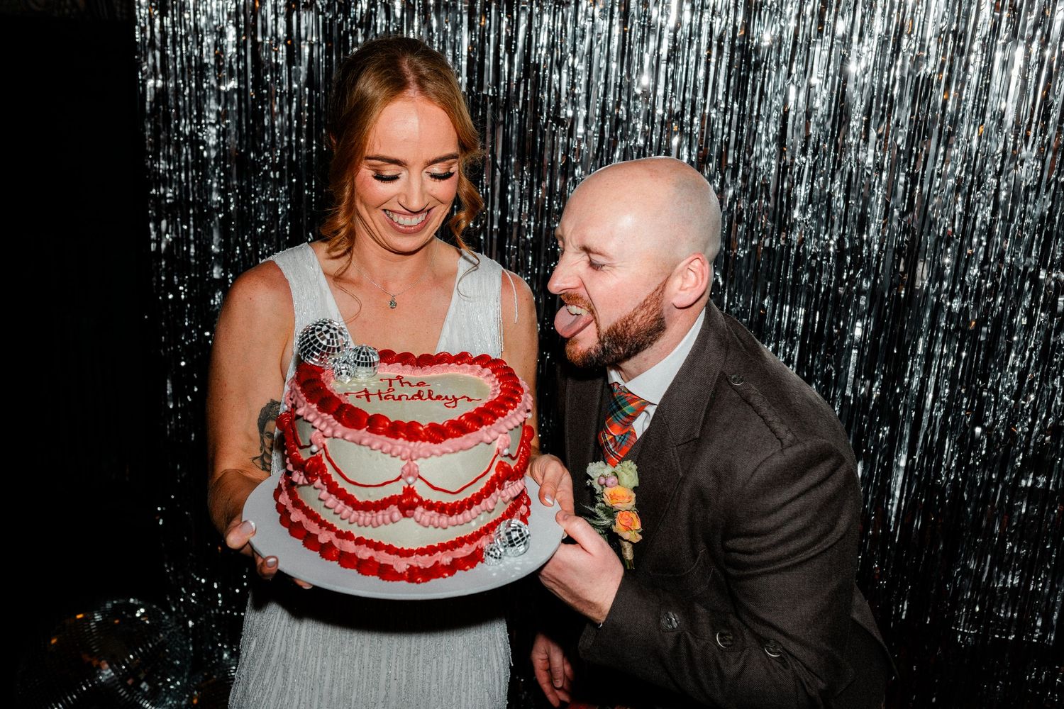 Bride and groom smiling while holding a decorated red birthday-style cake at their wedding reception.