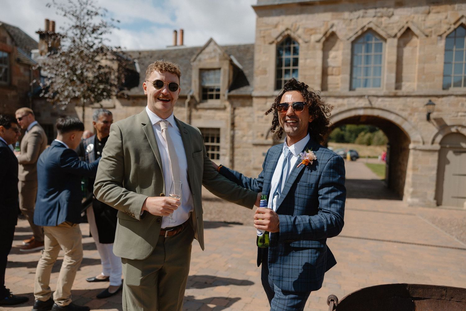 Two men in suits smiling at an outdoor wedding reception in a historic stone courtyard on a sunny day.